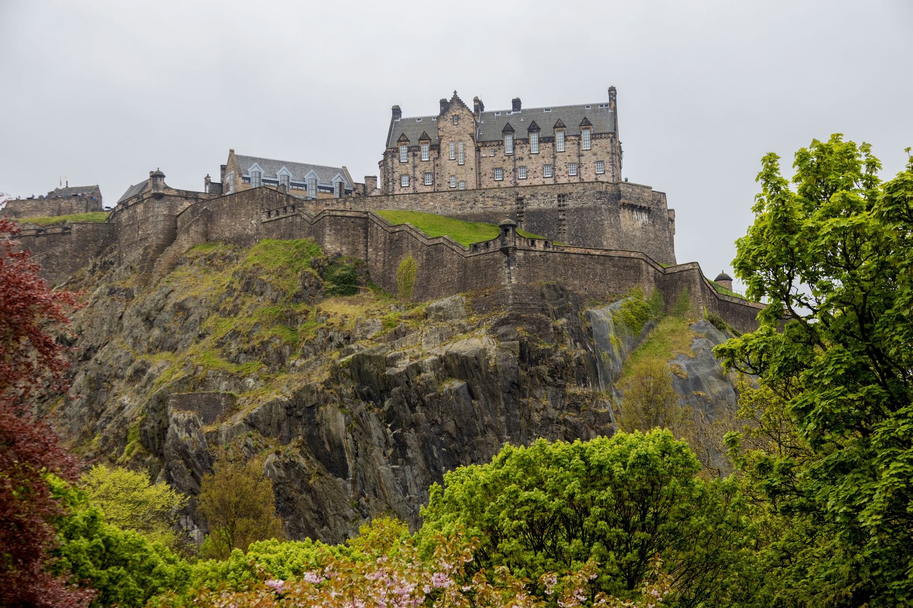 Il Castello di Edimburgo arroccato su una collina rocciosa, circondato da una rigogliosa vegetazione sotto un cielo nuvoloso.