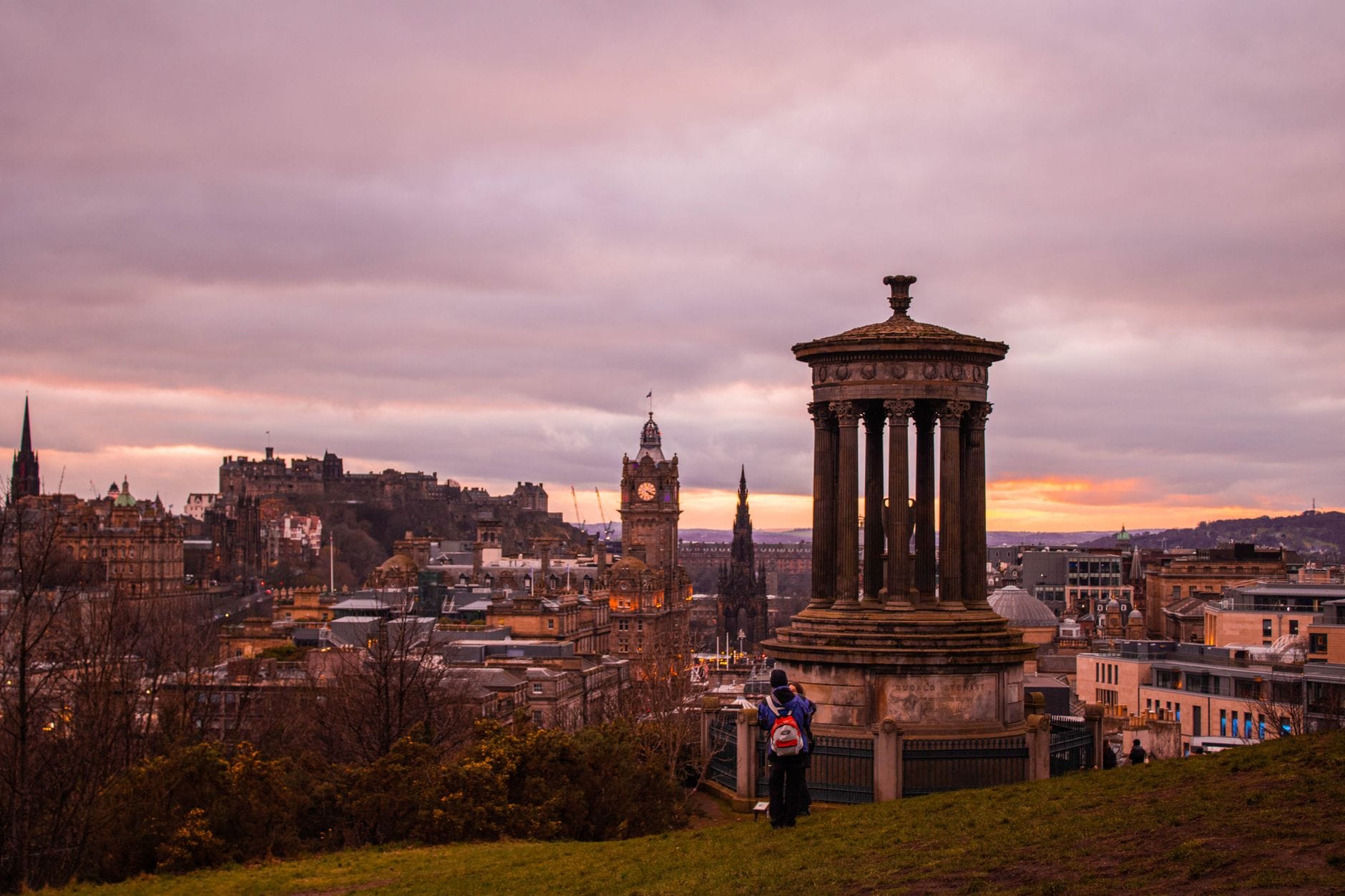 Panoramablick auf Edinburgh vom Calton Hill bei Sonnenuntergang, mit dem Dugald-Stewart-Monument, Stadtdächern und dem Edinburgh Castle im Hintergrund.
