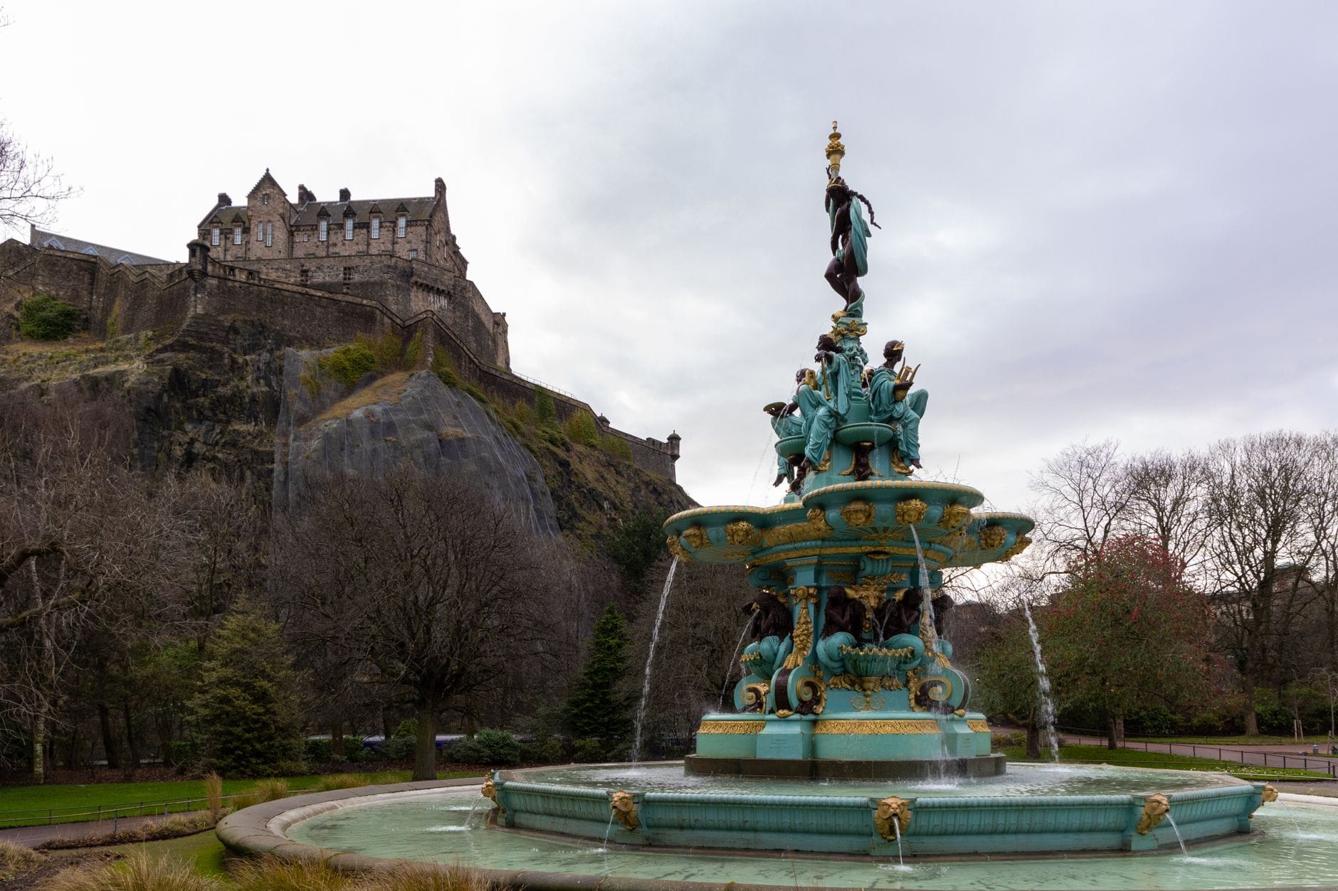 Vista della Ross Fountain nei Princes Street Gardens con l'Edinburgh Castle sulla collina sullo sfondo, circondato da alberi e aree verdi.