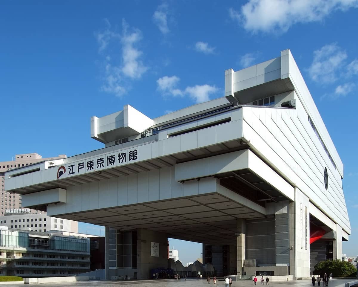 The futuristic architecture of the Edo-Tokyo Museum in Ryogoku stands against a bright blue sky, with visitors walking below the raised structure.