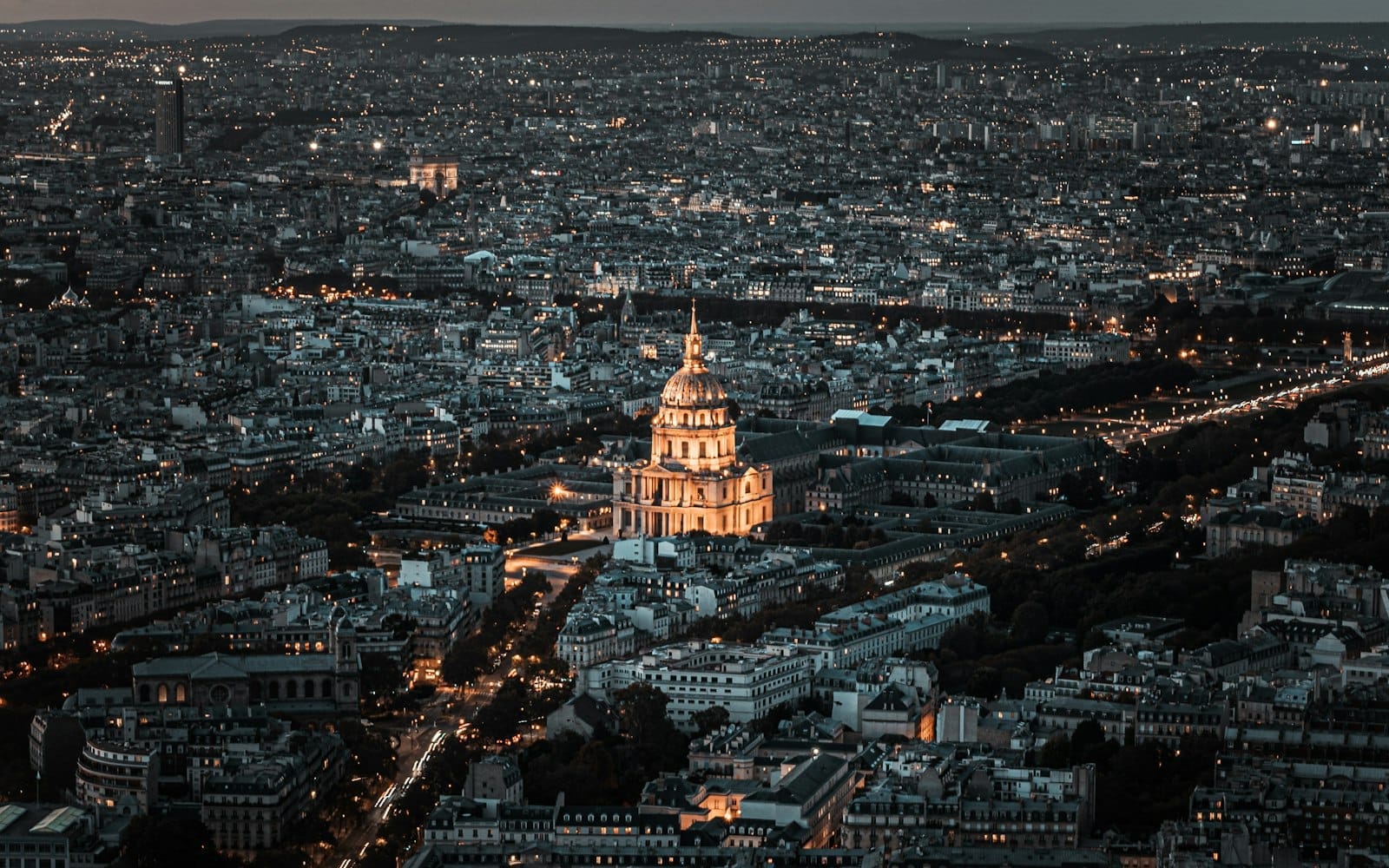 Vista aerea di Parigi al tramonto con la cupola dorata illuminata di Les Invalides che spicca al centro, mentre il panorama urbano si estende fino all'orizzonte.