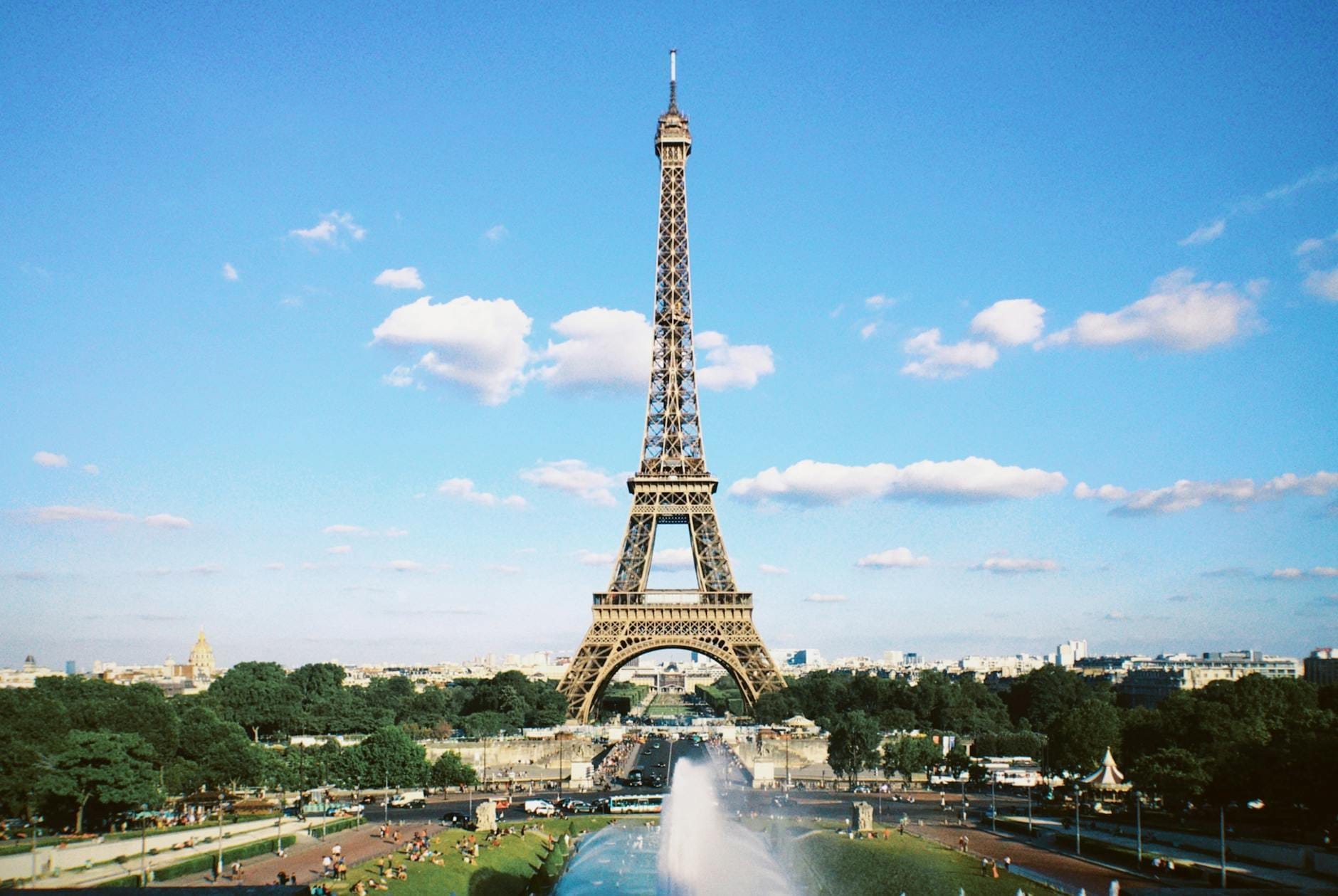Vista ampia della Torre Eiffel su un cielo blu, con giardini verdi, una fontana e persone che godono dell'atmosfera parigina.