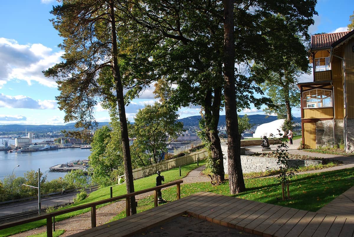 วิวของสวนประติมากรรม Ekebergparken พร้อมต้นไม้สีเขียว เส้นทางเดิน ประติมากรรม และ Oslofjord ที่มองเห็นได้ภายใต้ท้องฟ้าสีฟ้าที่มีเมฆกระจาย