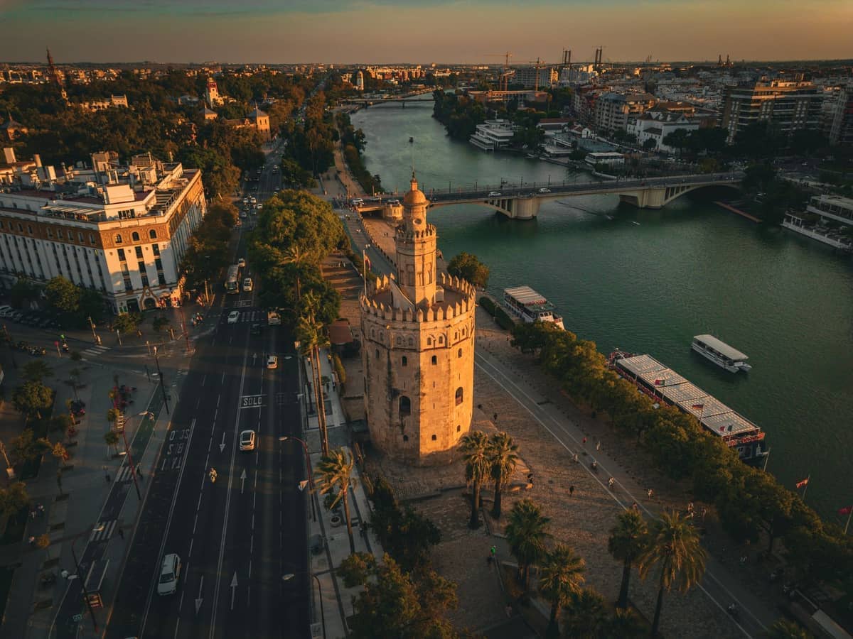 มุมมองทางอากาศของ El Arenal ในเมือง Seville ที่มี Torre del Oro อันเป็นสัญลักษณ์ ทางเดินริมแม่น้ำ และเรือบน Guadalquivir ยามพระอาทิตย์ตกดิน