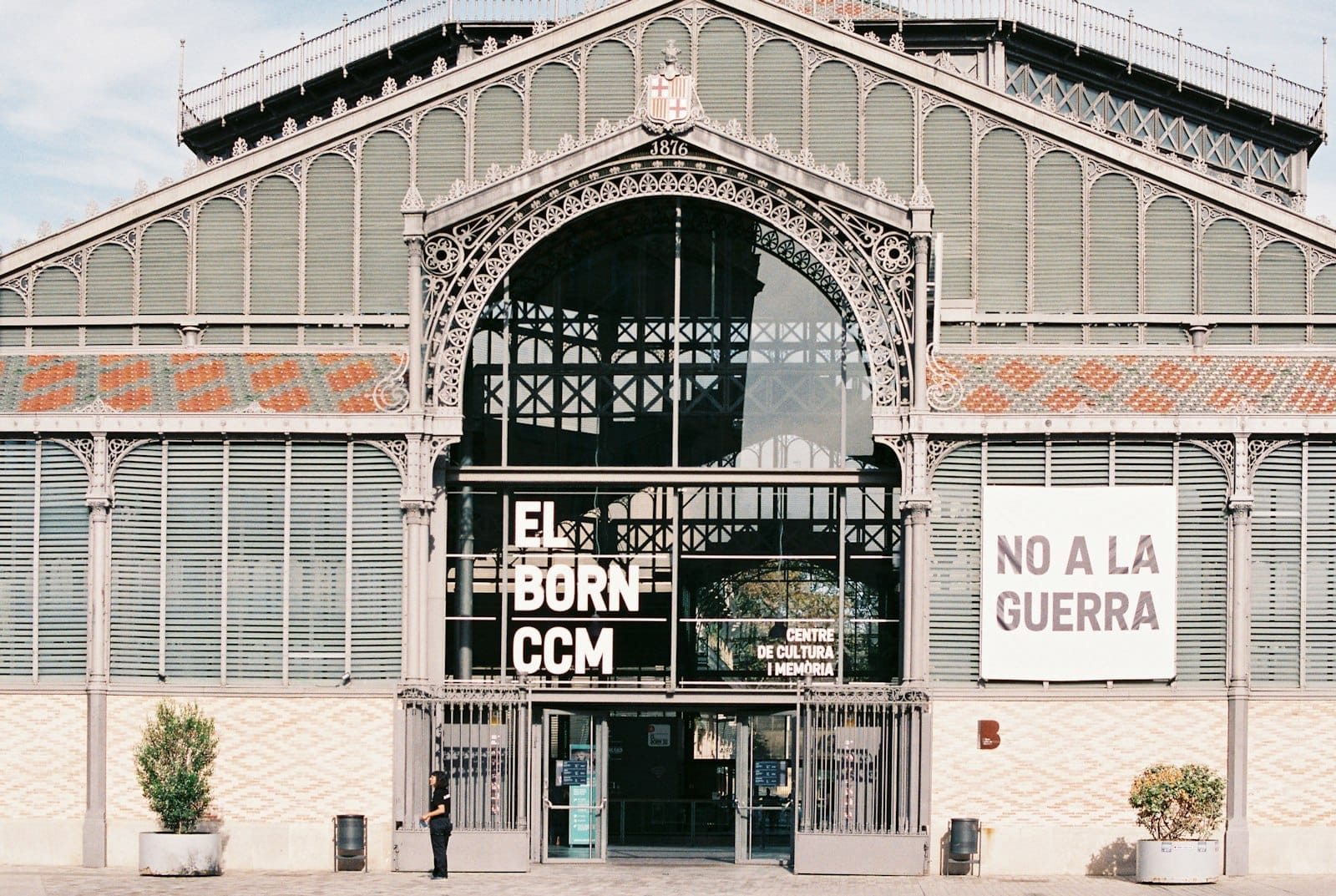 Front view of El Born Centre de Cultura i Memòria, featuring its ornate iron architecture, banners, and a single person by the entrance.