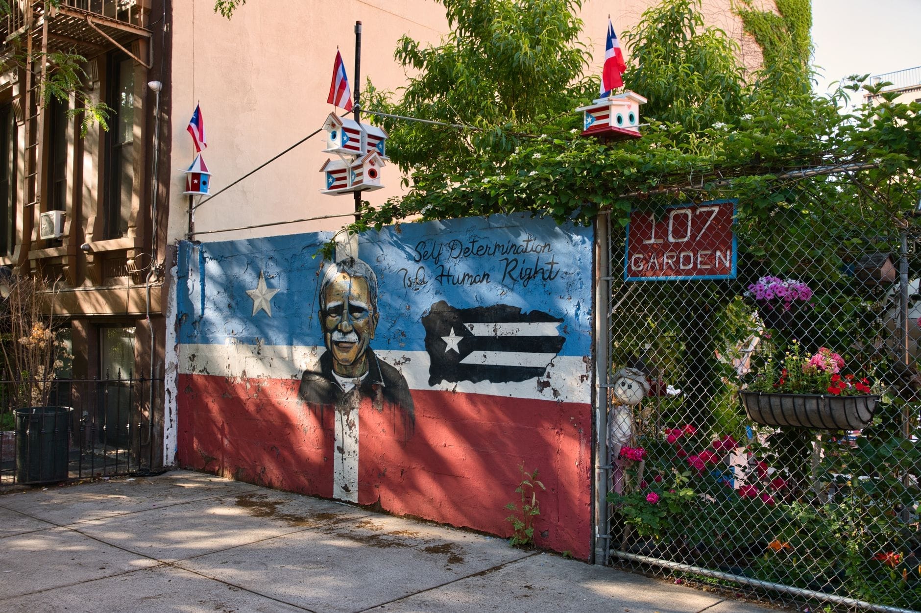 Murale colorato con un ritratto e la bandiera portoricana in un giardino comunitario di East Harlem, decorato con piccole bandierine e fiori in una giornata di sole.
