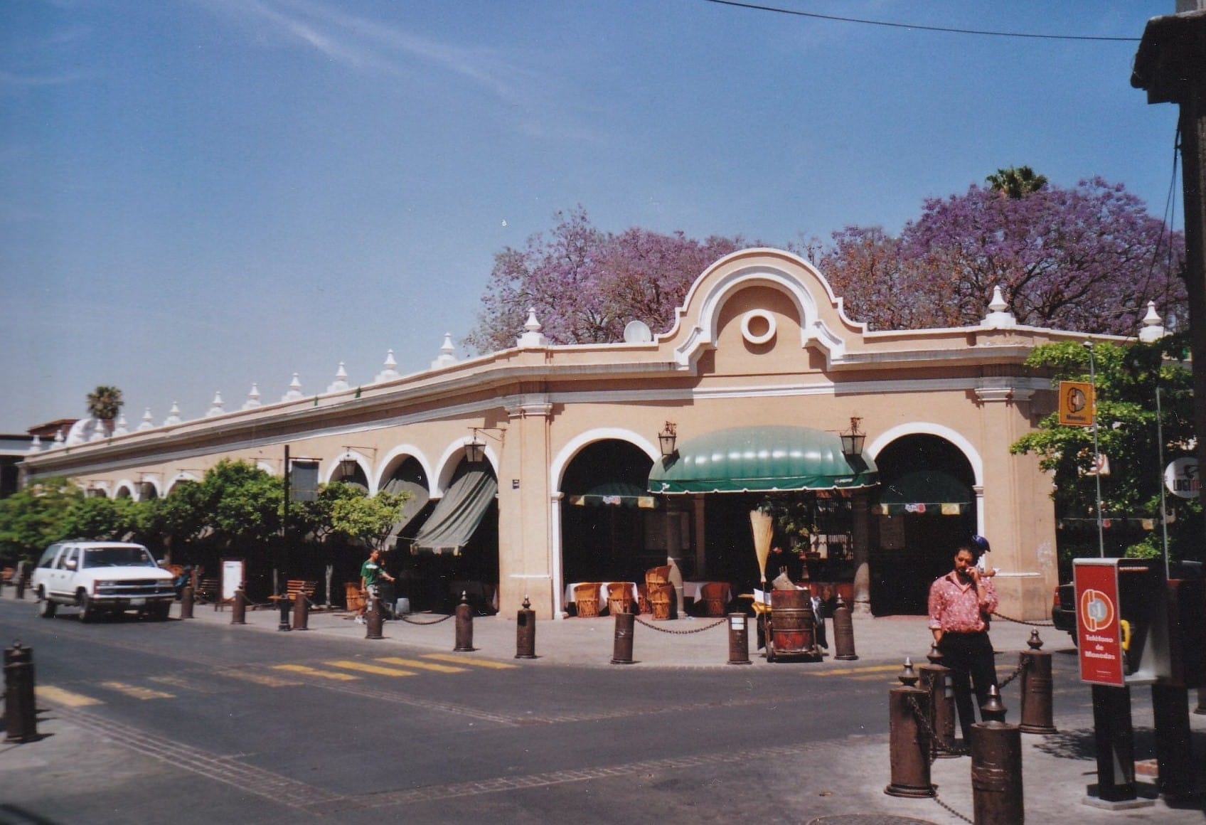 Lo storico El Parián de Tlaquepaque con facciate ad archi e tendoni verdi, circondato da jacaranda in fiore sotto un cielo azzurro limpido.