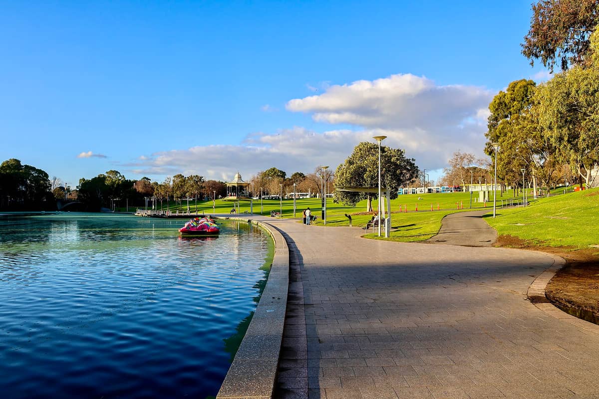 Klarer Blick auf den Elder Park mit dem River Torrens, geschwungenem Weg, grünen Wiesen, Pavillon und Tretbooten unter strahlend blauem Himmel in Adelaide.