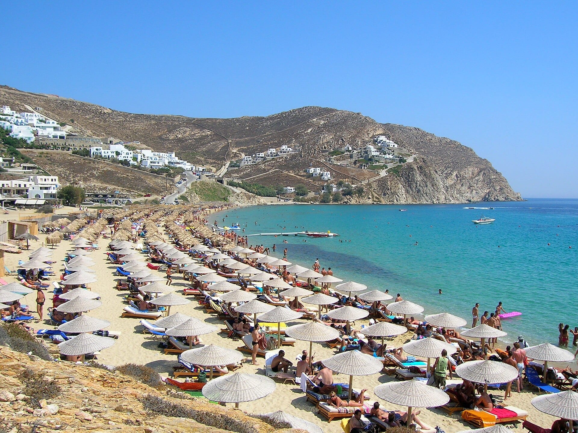 Hileras de tumbonas y sombrillas bordean la playa de Elia en Mykonos, con aguas turquesas y edificios blancos en la ladera bajo un cielo azul despejado.