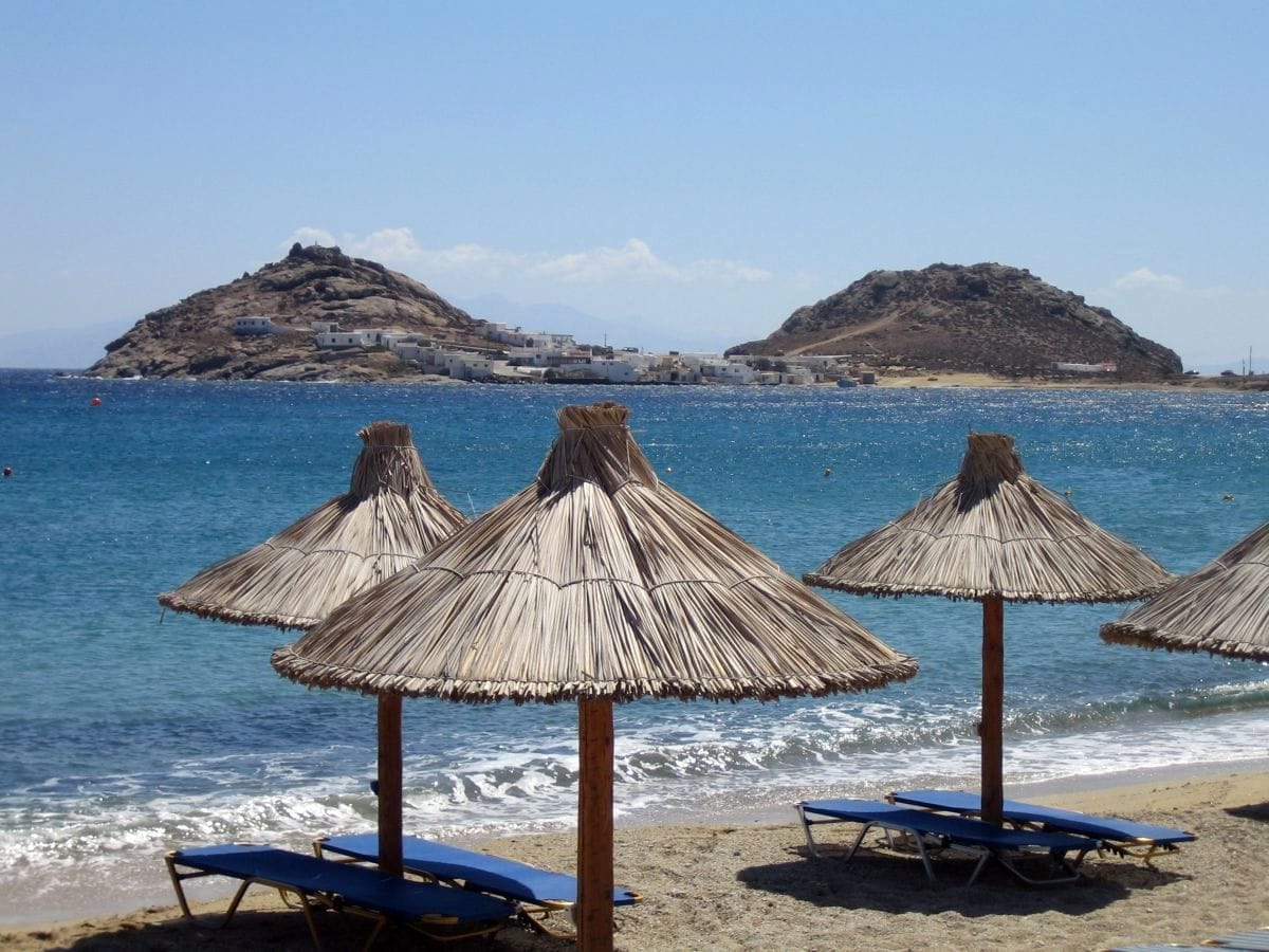 Sombrillas de playa con techo de paja y tumbonas azules en Elia Beach, Mykonos, con agua turquesa cristalina y colinas rocosas al fondo.