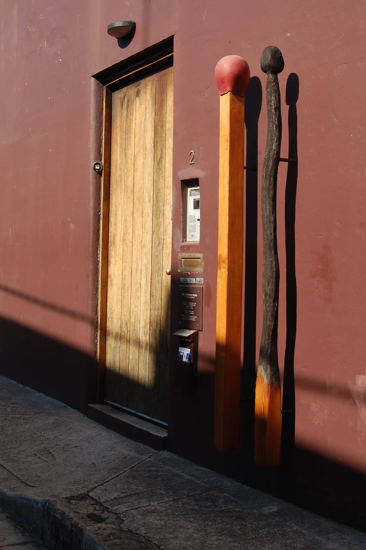 Puerta de madera en una pared color burdeos, flanqueada por dos esculturas gigantes de cerillas que proyectan largas sombras bajo la luz del sol de la tarde.