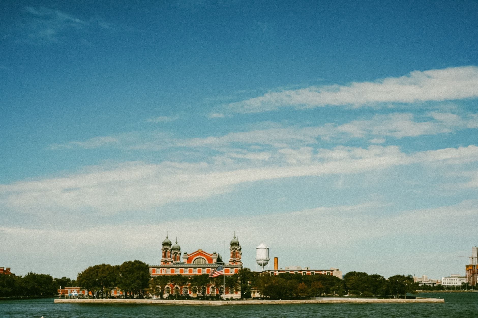 Vista panoramica dell'edificio principale Beaux-Arts di Ellis Island circondato da alberi, con l'acqua e un cielo blu striato di nuvole in primo piano.