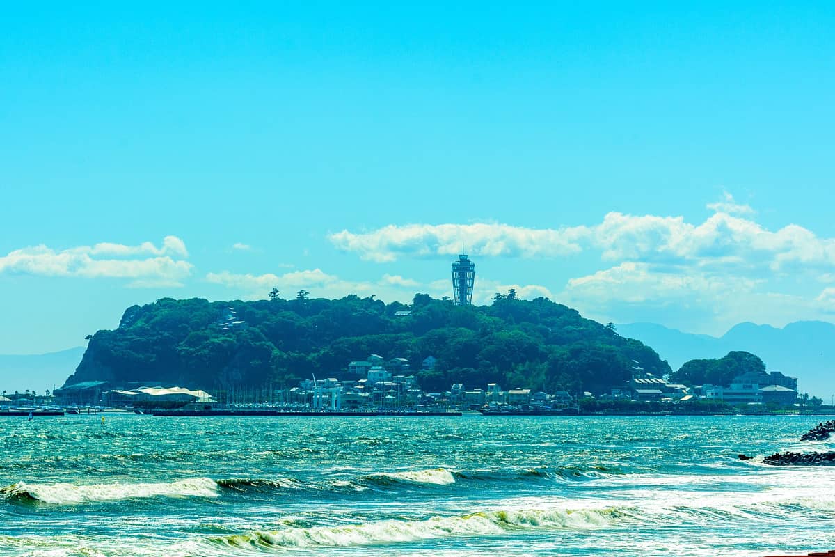 Distant view of Enoshima Island rising from the sea, with its iconic observation tower visible against a blue sky and mountains in the background.
