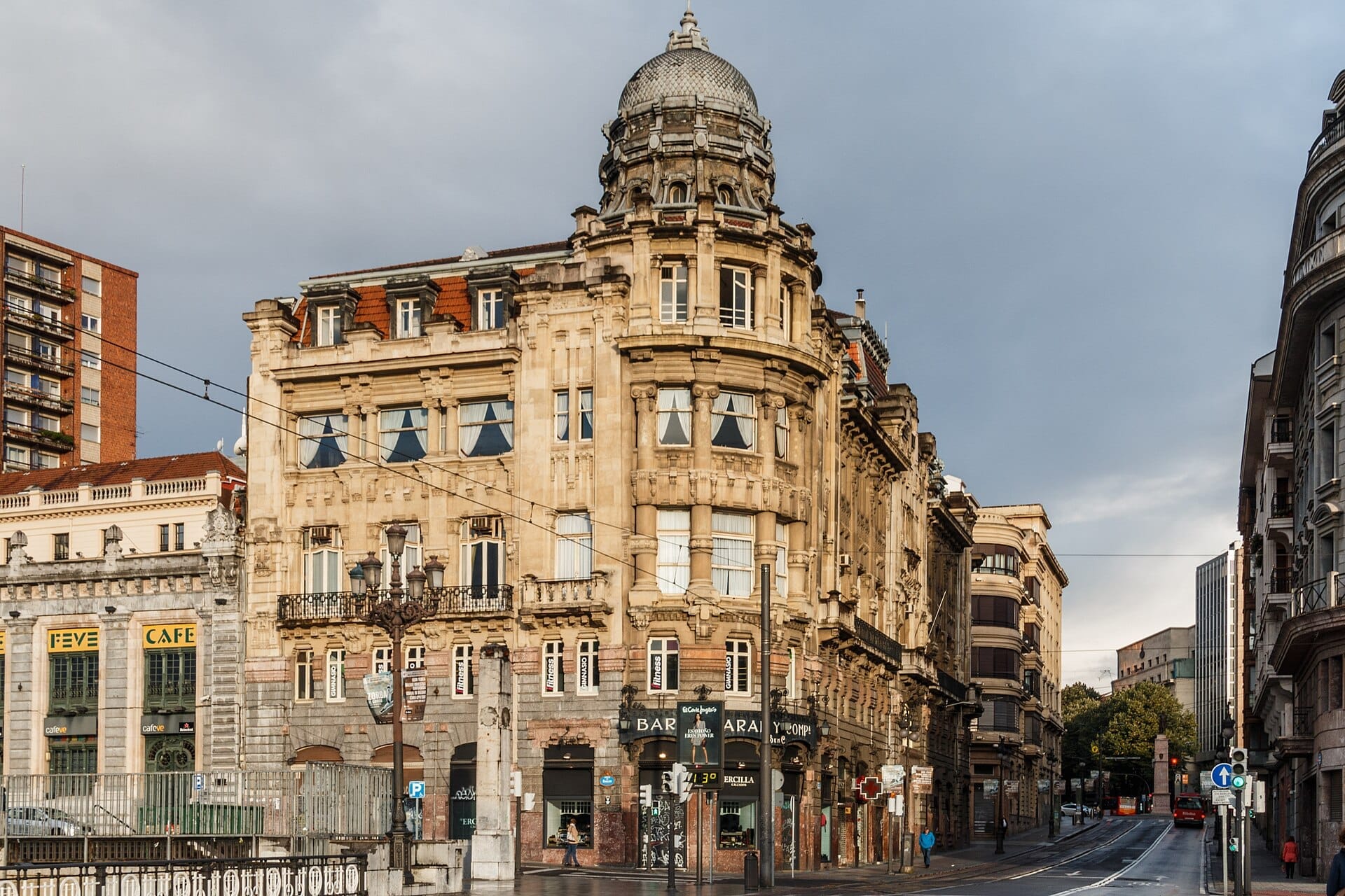 Classic architecture with ornate facades and a domed rooftop stands at a street intersection in Bilbao’s Ensanche district on a cloudy day.