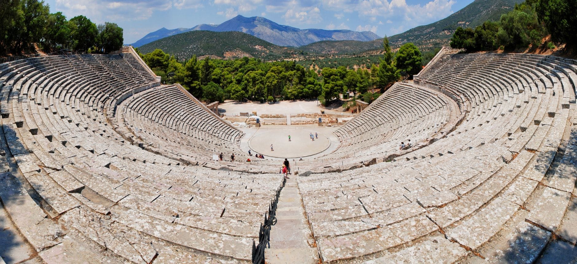 Vista panoramica del Teatro Antico di Epidauro con le ampie gradinate in pietra, circondato da una rigogliosa vegetazione verde e montagne lontane sotto un cielo azzurro.