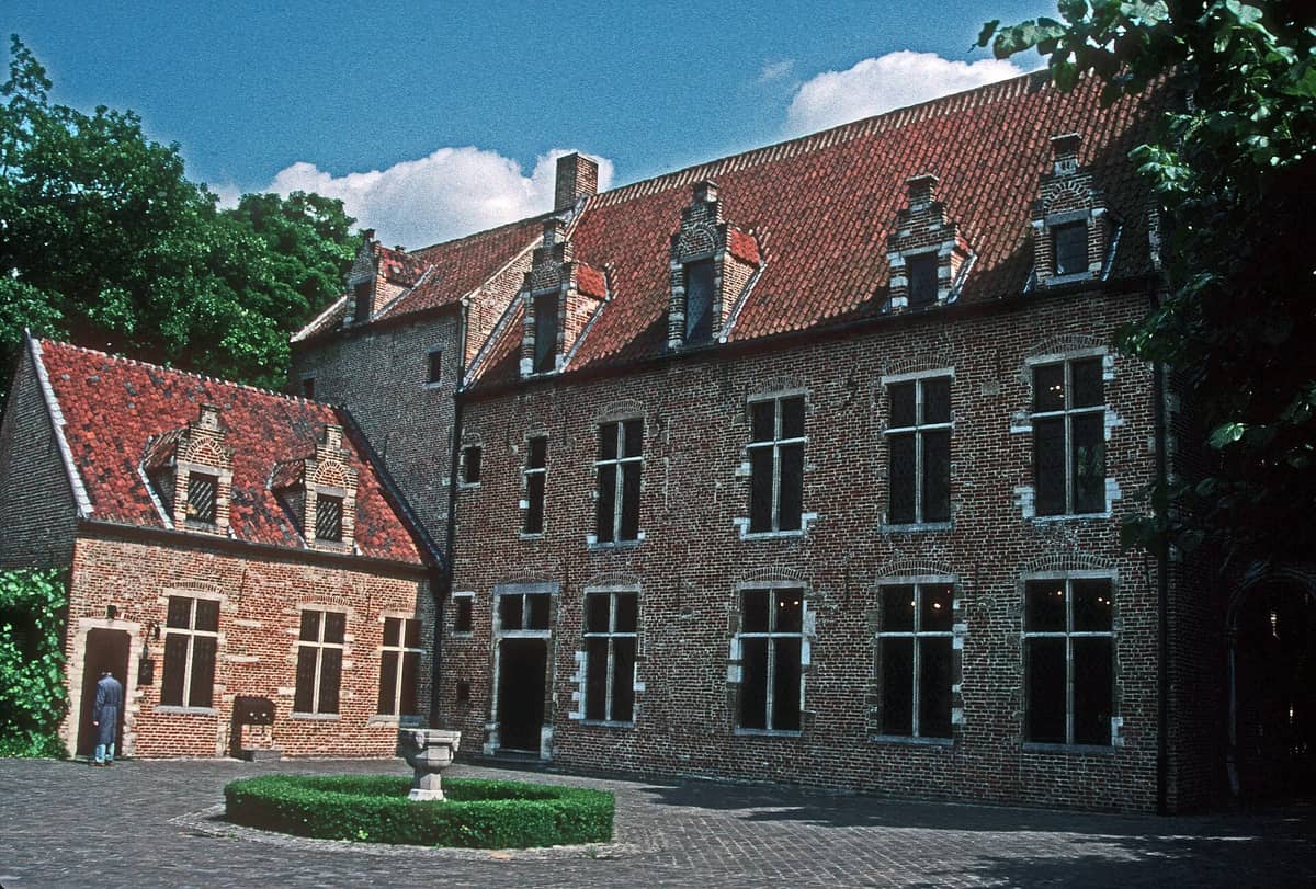 Vista exterior da Casa de Erasmo, em tijolos vermelhos, com janelas gótico-tardias, telhado íngreme de telhas e vegetação exuberante sob um céu azul em Anderlecht, Bruxelas.