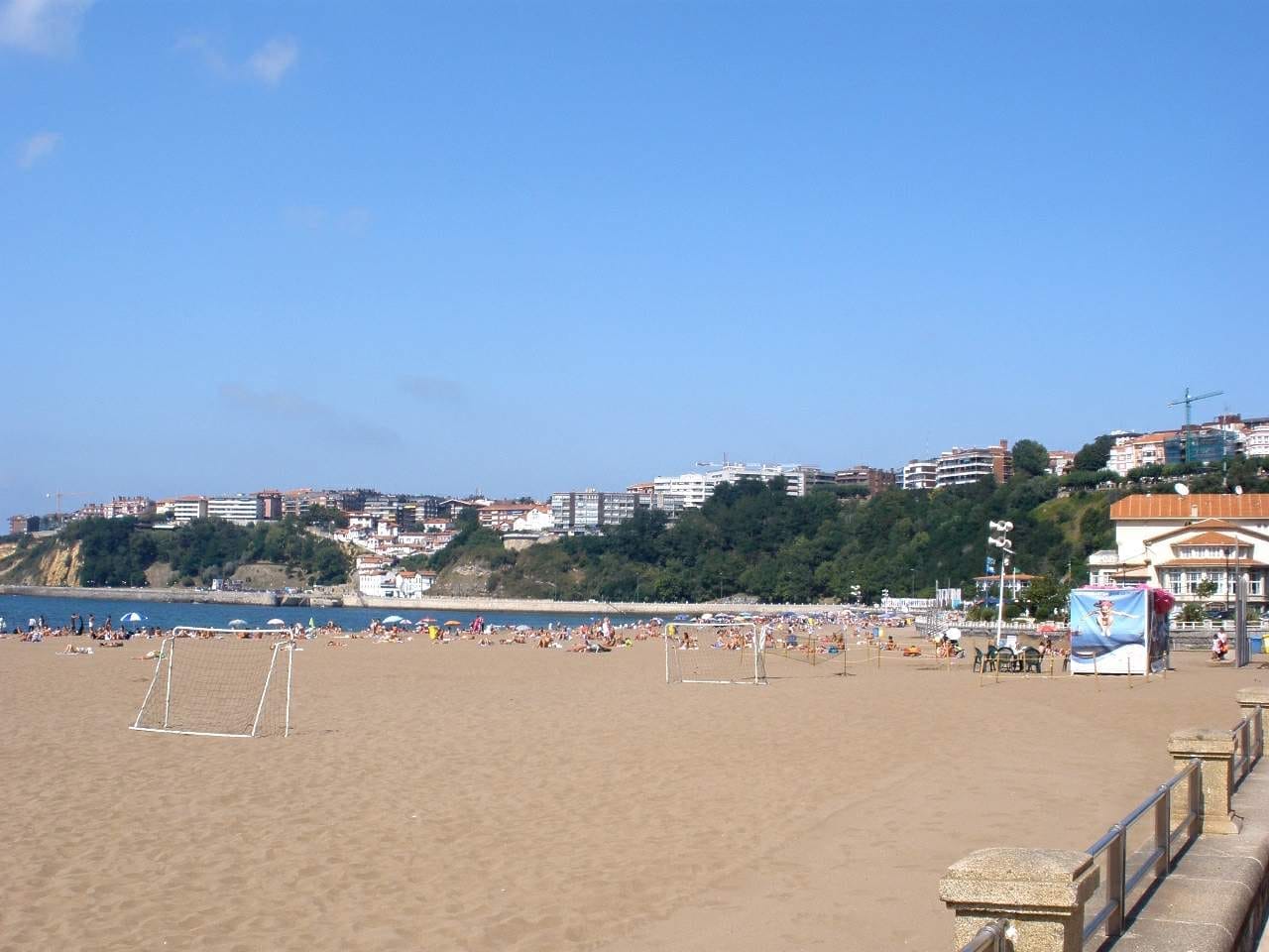 Blick auf den Strand Ereaga in Getxo mit goldenem Sand, vereinzelten Sonnenbadenden, fernem Meer, städtischen Gebäuden und grünen Hügeln unter einem klaren blauen Himmel.
