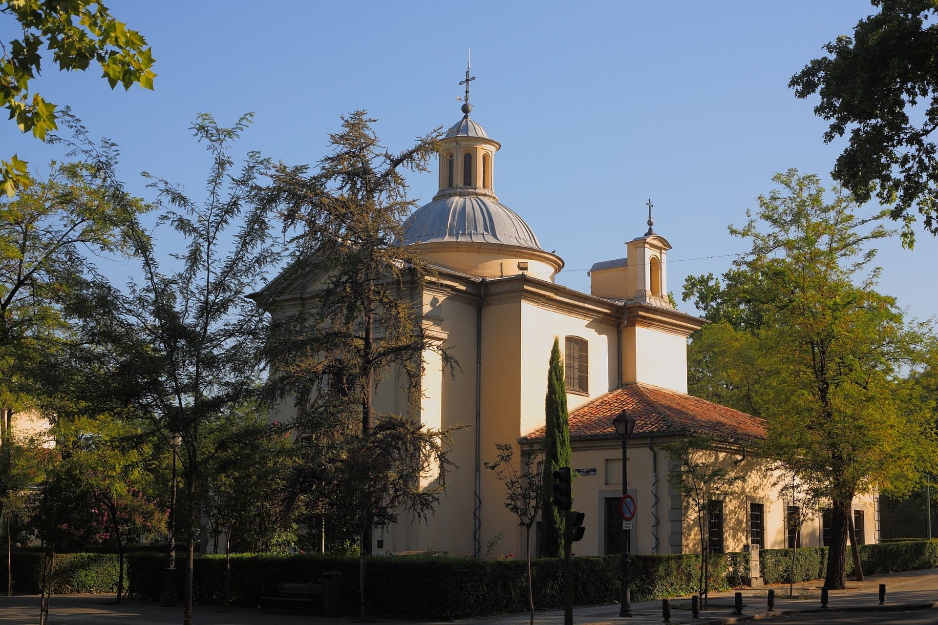 Vista dell'Ermita de San Antonio de la Florida con la sua cupola neoclassica, la facciata giallo chiaro e gli alberi verdi circostanti in una giornata serena.