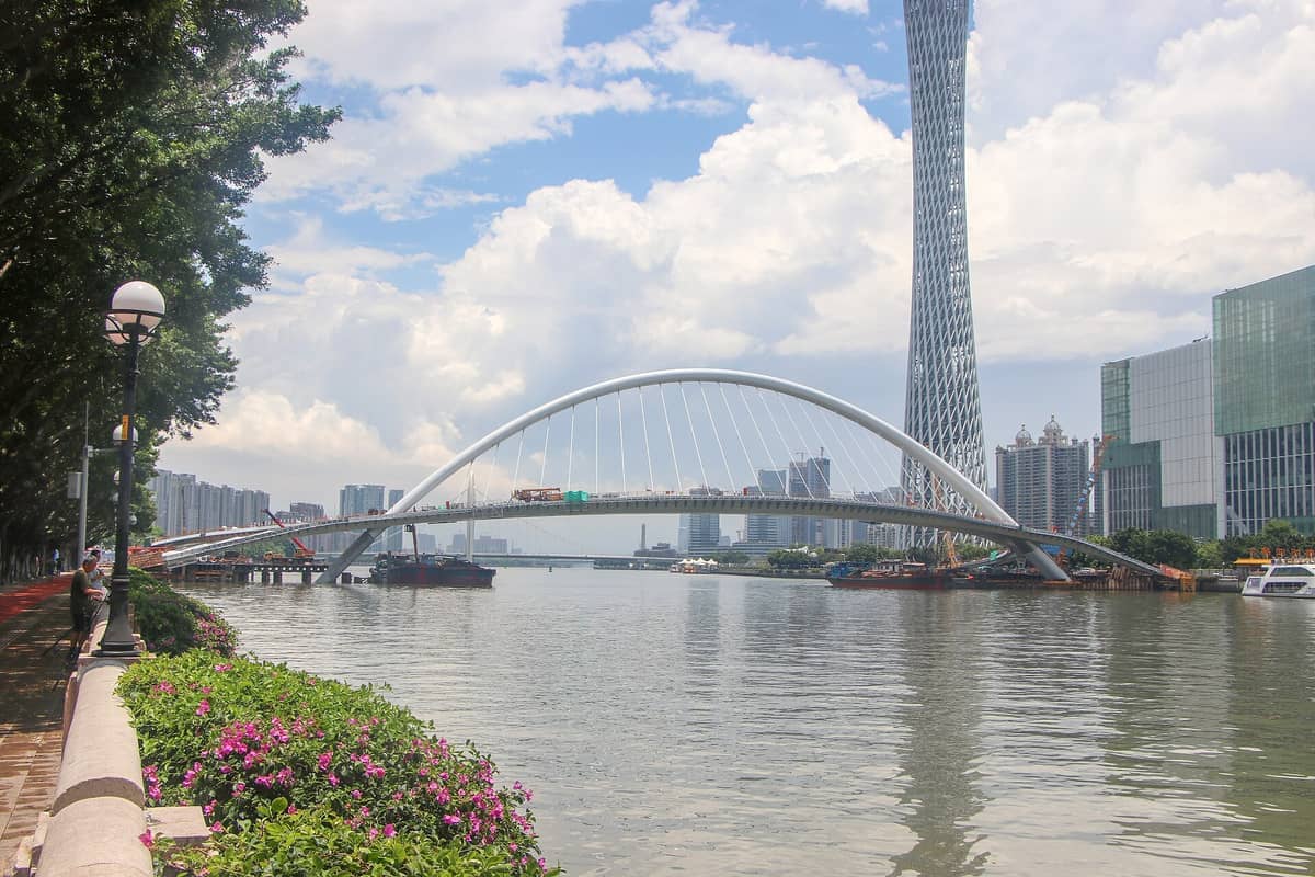 View of Ersha Island with lush riverside walkway, blooming pink flowers, landmark bridge over the Pearl River, and Guangzhou skyline under a bright sky.