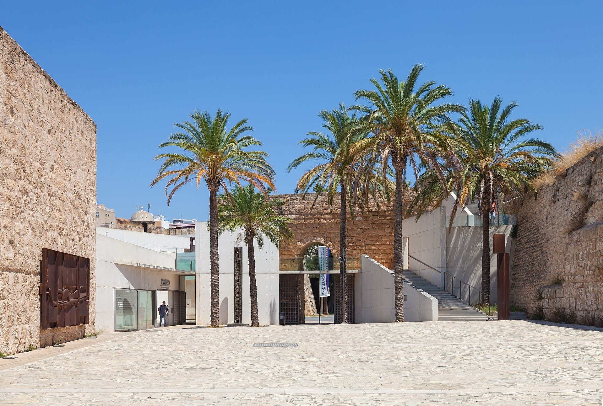 Cortile d'ingresso del Museo Es Baluard a Palma, con muri in pietra, architettura moderna, palme e cielo blu limpido.