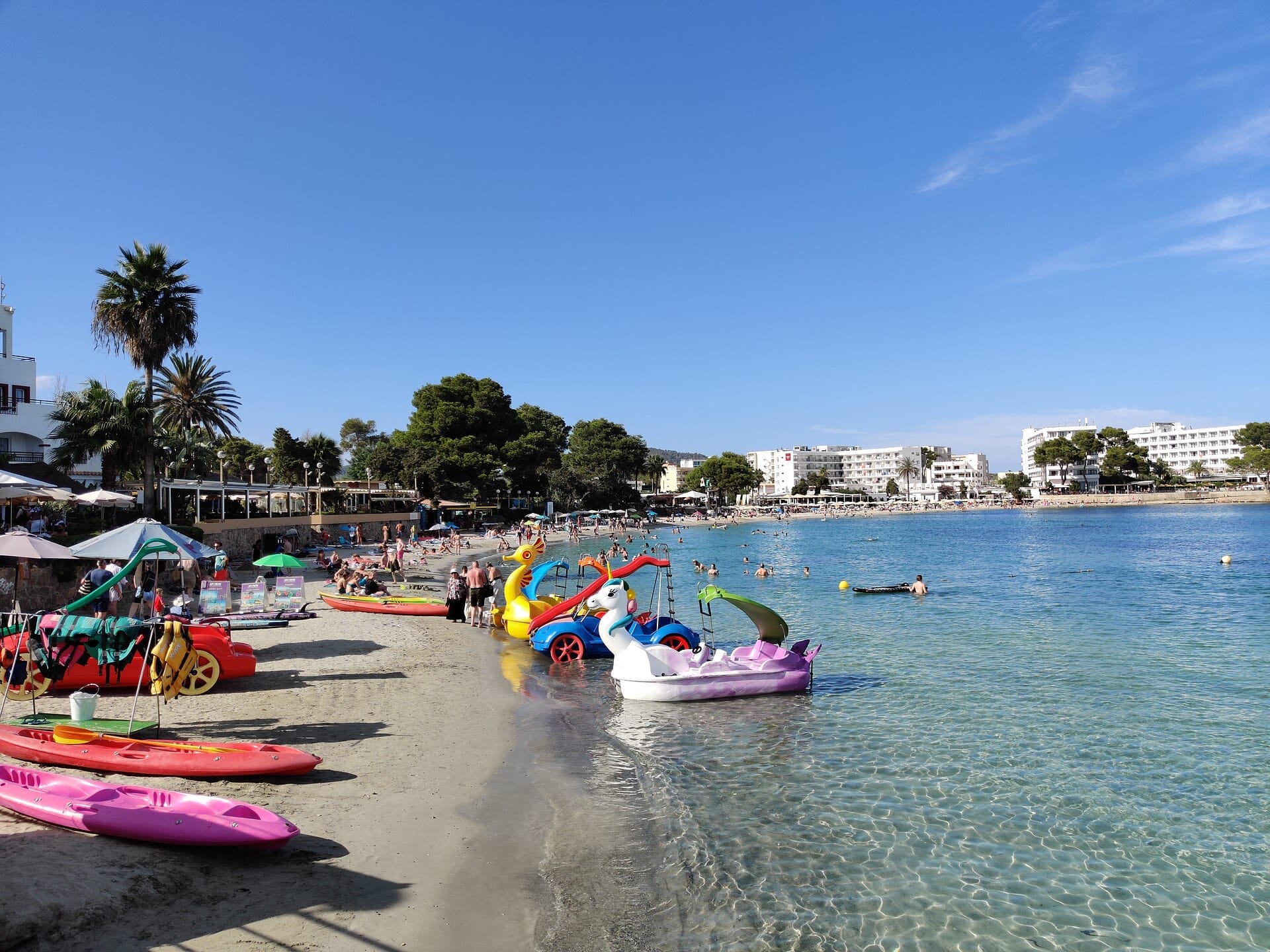 Kayak colorati e pedalò costeggiano la riva sabbiosa di Es Canar Beach, con bagnanti e nuotatori che godono delle limpide acque del Mediterraneo.