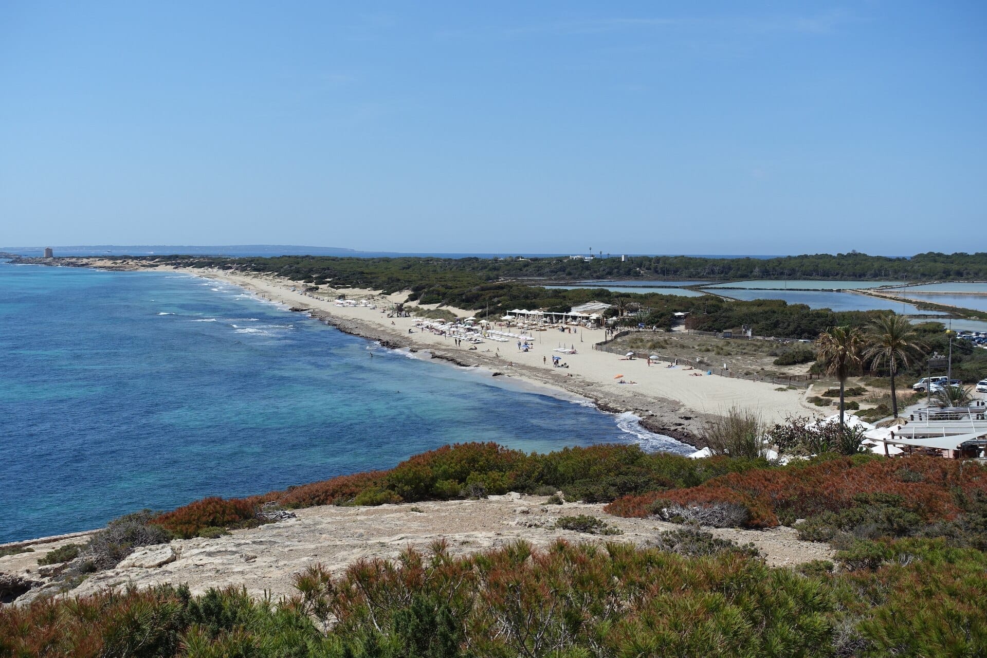 Vista aerea della spiaggia Es Cavallet che mostra la sua ampia distesa di sabbia bianca e morbida, acque turchesi cristalline, vegetazione costiera e le vicine saline.