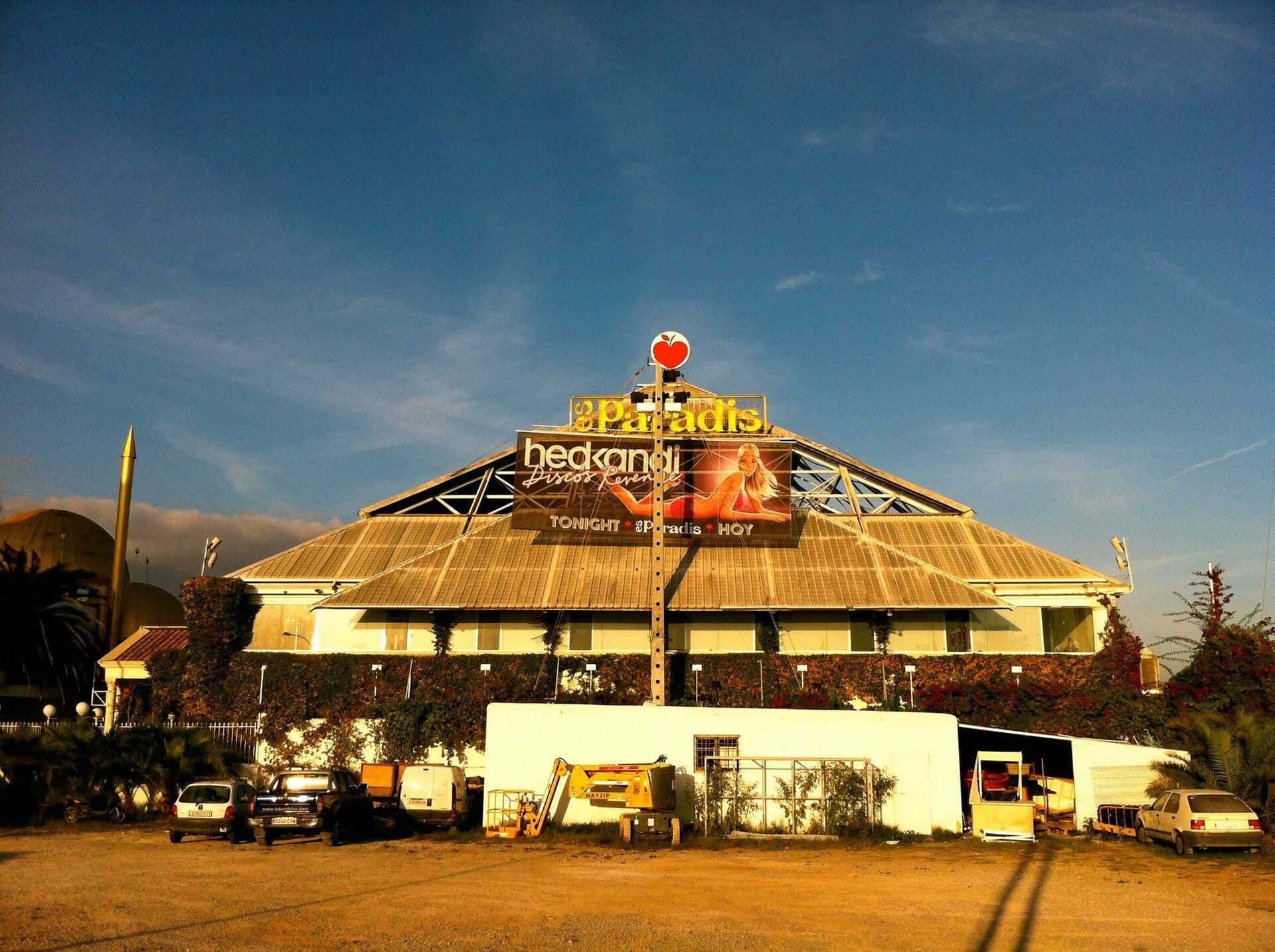 Vista esterna del nightclub Es Paradis a Ibiza, con il suo iconico tetto a piramide di vetro, grandi striscioni per eventi e veicoli parcheggiati sotto un cielo azzurro limpido.