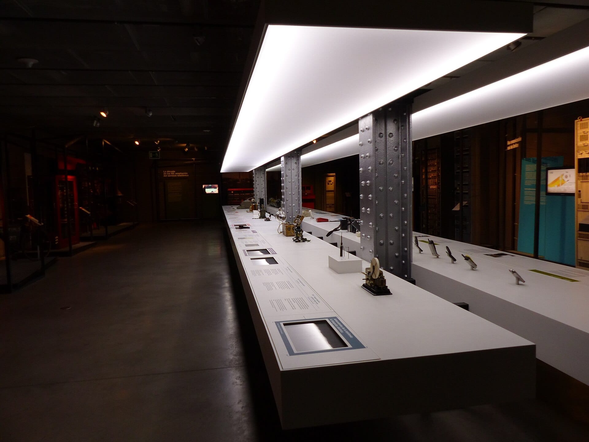 Galleria espositiva moderna all'Espacio Fundación Telefónica con tavoli espositivi bianchi illuminati, travi industriali e manufatti tecnologici storici sotto un'illuminazione ambientale.