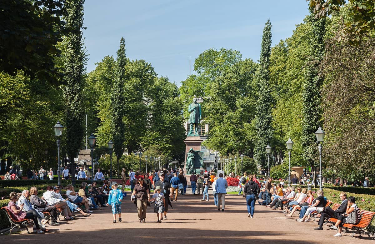 Pessoas relaxando nos bancos e caminhando pelo caminho principal arborizado do Parque Esplanadi no verão, com a estátua de Johan Ludvig Runeberg visível ao fundo.