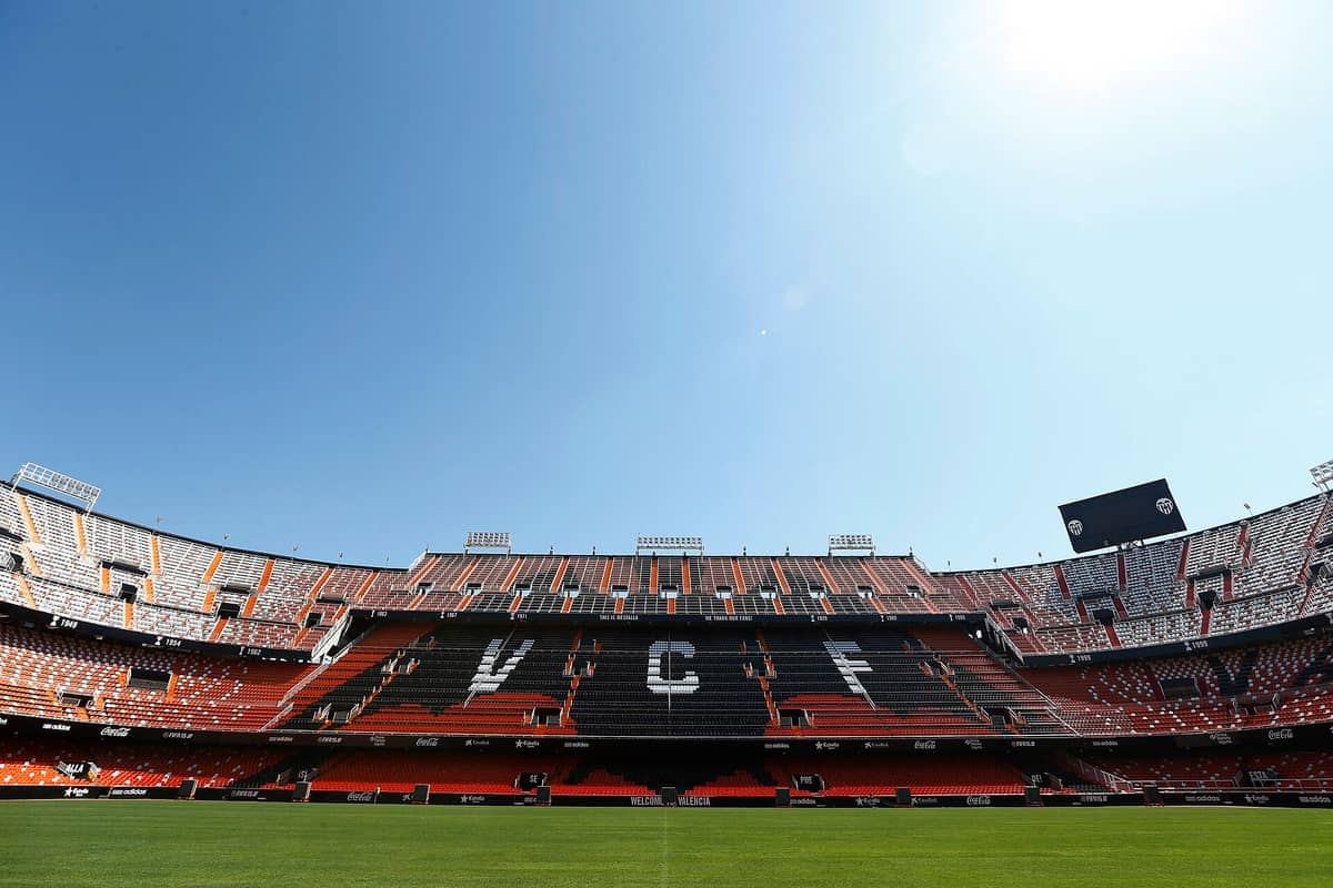 Vista grandangolare delle tribune vuote rosse e bianche dell'Estadio de Mestalla con la scritta VCF, sotto un cielo azzurro brillante.