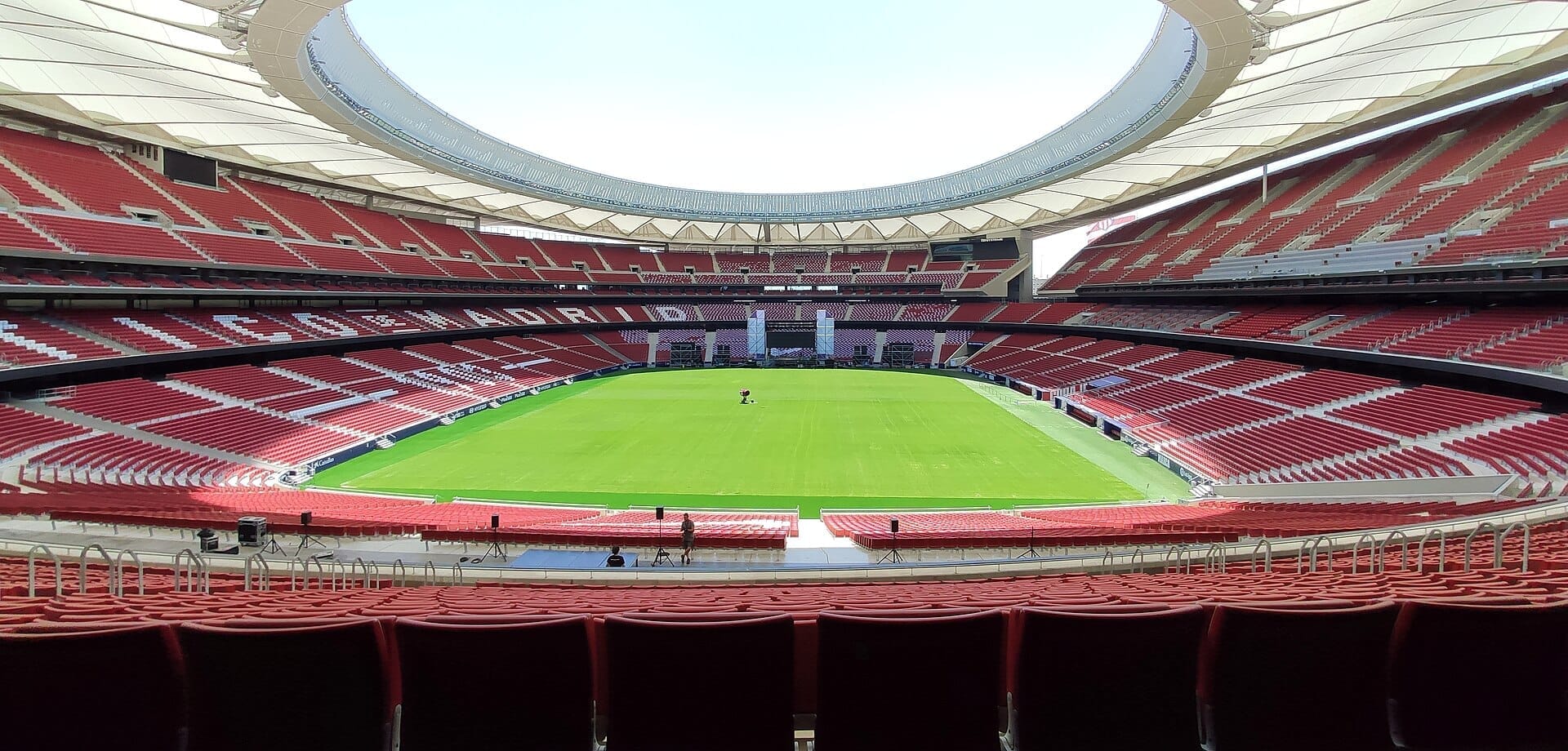 Vista ampia dello stadio Riyadh Air Metropolitano con sedute rosse, tribune vuote e un manto erboso verde ben curato sotto un cielo luminoso.