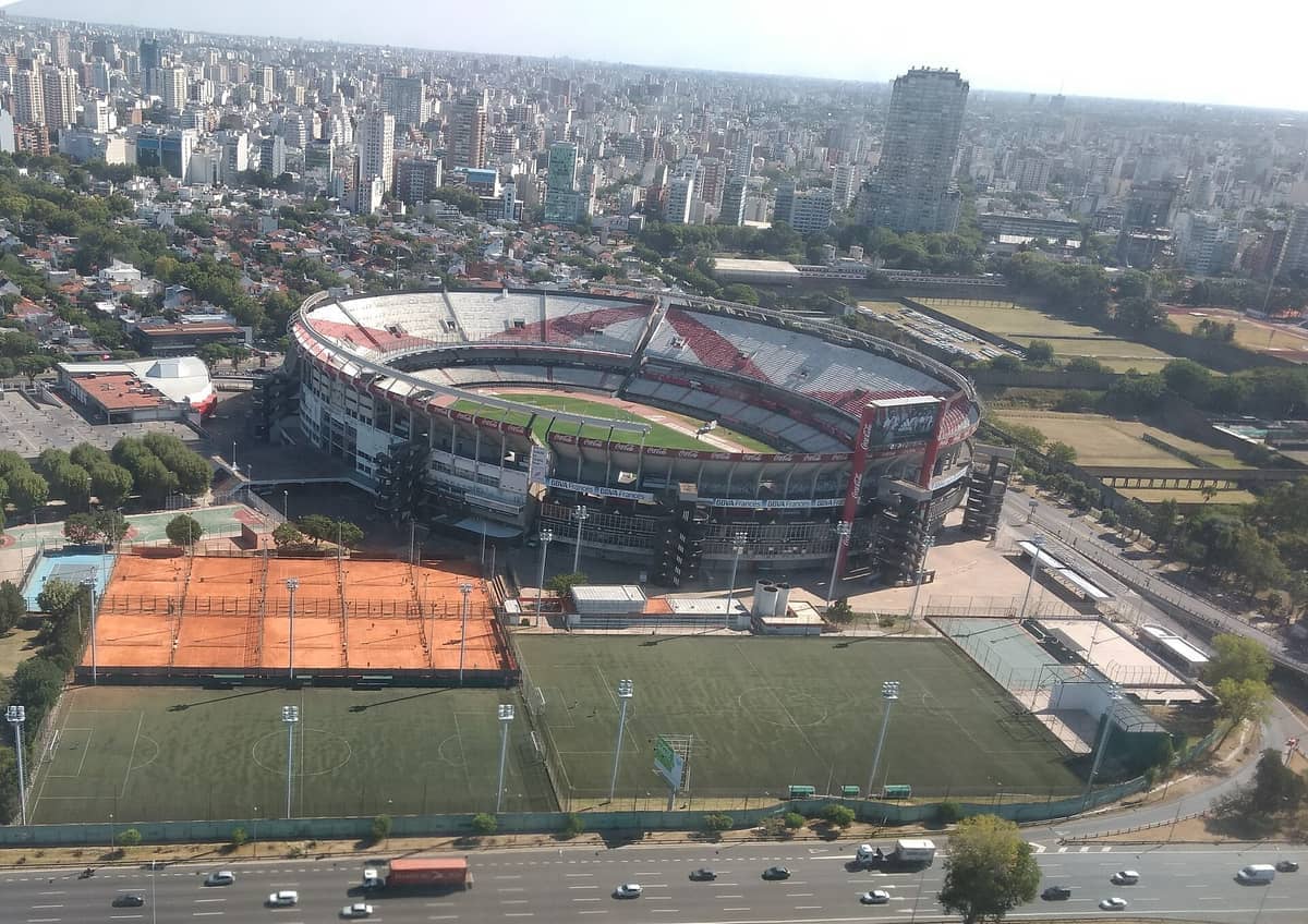 มุมมองทางอากาศของ Estadio Más Monumental ในบัวโนสไอเรส ล้อมรอบด้วยอาคารในเมือง สนามหญ้าสีเขียว และสนามกีฬาที่อยู่ติดกันในวันที่อากาศแจ่มใส