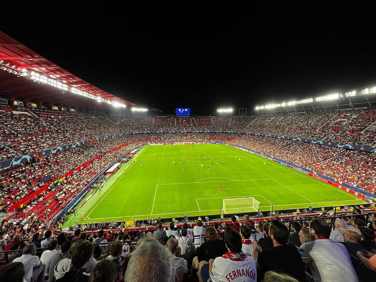 ภาพมุมกว้างของ Estadio Ramón Sánchez-Pizjuán ที่เต็มไปด้วยผู้ชมในยามค่ำคืน พร้อมแฟนบอลหลายพันคนที่กำลังชมการแข่งขันฟุตบอลภายใต้แสงไฟสนามที่สว่างจ้า