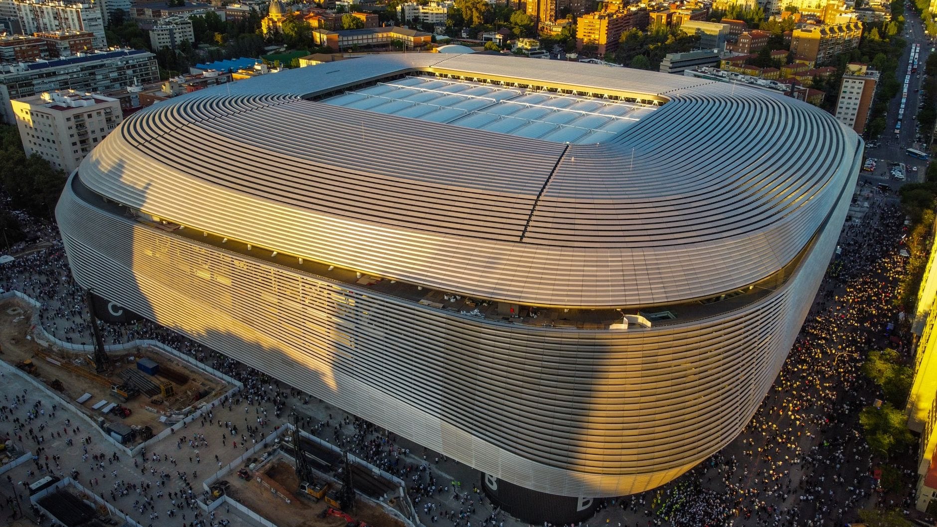Vista aerea al tramonto dell'Estadio Santiago Bernabéu ristrutturato, circondato dalla folla e dagli edifici della città, con la sua architettura moderna e la sua imponente grandiosità.