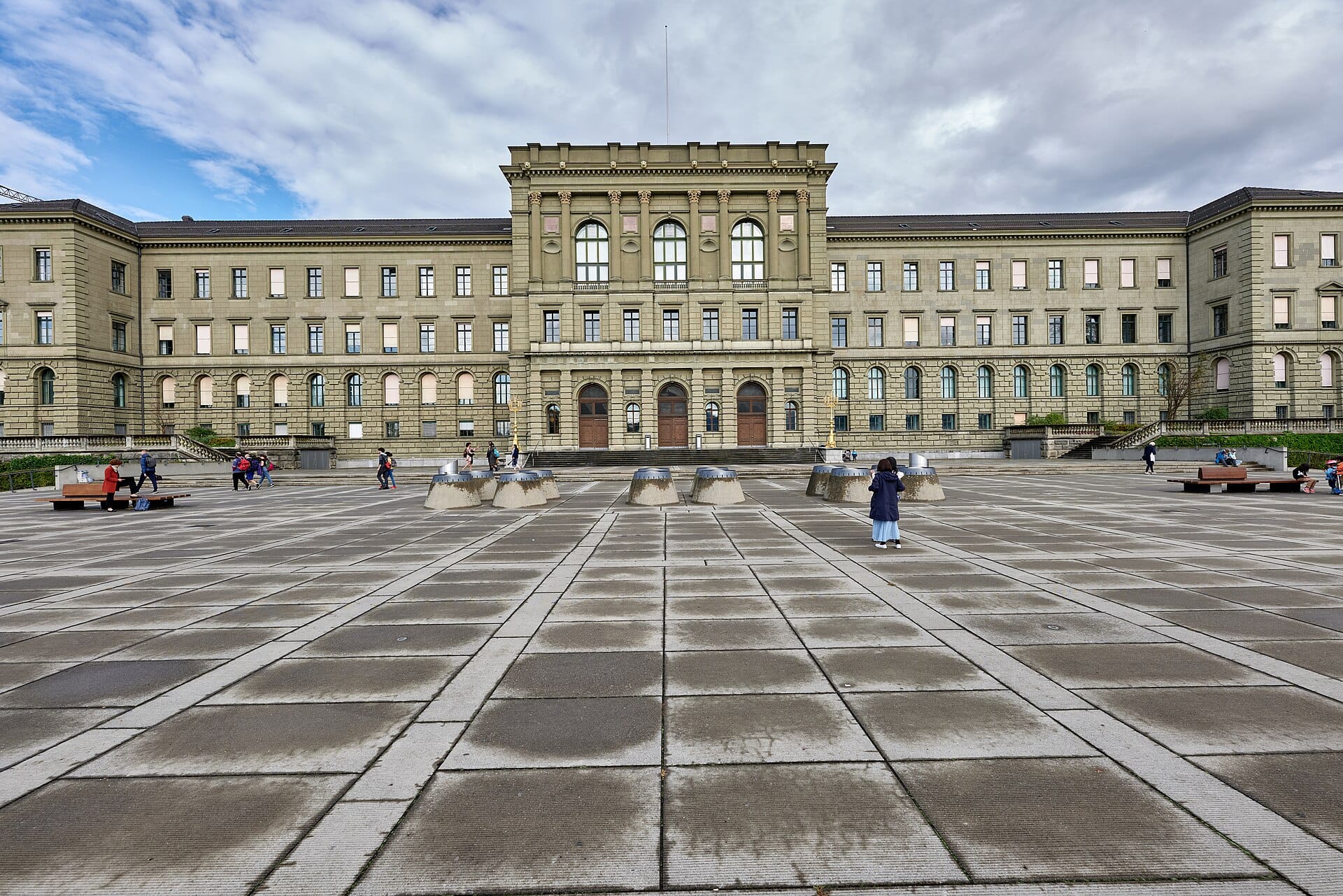 ภาพมุมกว้างด้านหน้าอาคารหลัก ETH Zurich โชว์สถาปัตยกรรมนีโอคลาสสิก พื้นที่จัตุรัสหลัก มีผู้คน ม้านั่ง และหน้าต่างโค้ง ภายใต้ท้องฟ้าครึ้มบางส่วน