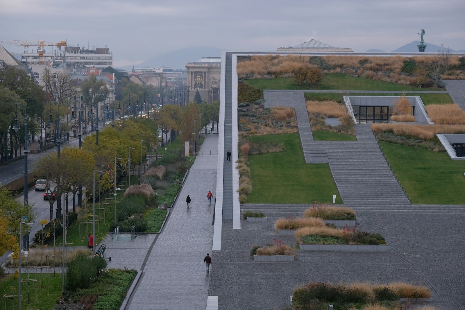 Moderner Architekturbau des Völkerkundemuseums in Budapest mit geometrischen Dachgärten und Besuchern, die eine breite, begrünte Allee entlanggehen.