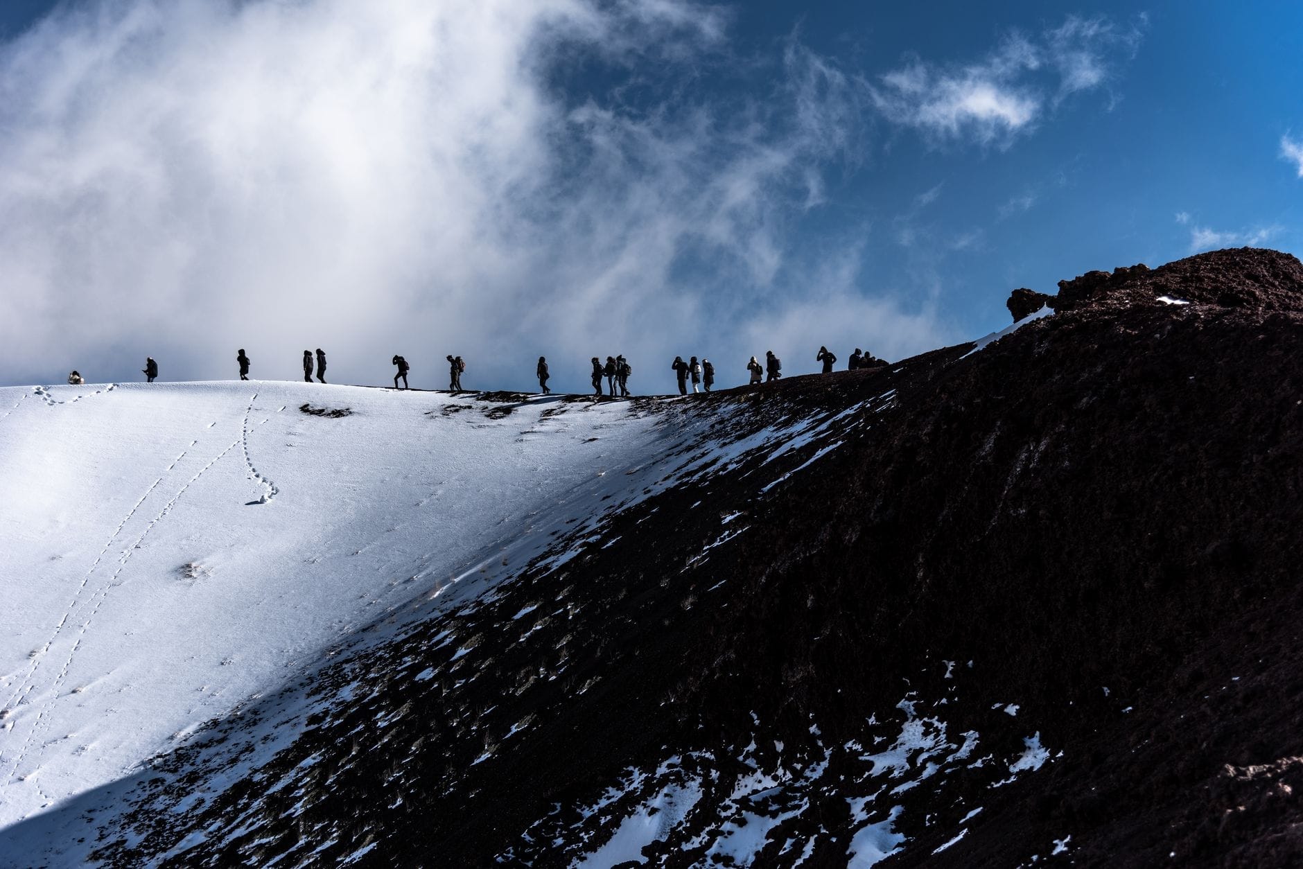 Eine Gruppe von Wanderern läuft entlang eines verschneiten Grats nahe dem Gipfel des Ätna, unter dramatischen Wolken und blauem Himmel – Abenteuer und Vulkanlandschaft pur.
