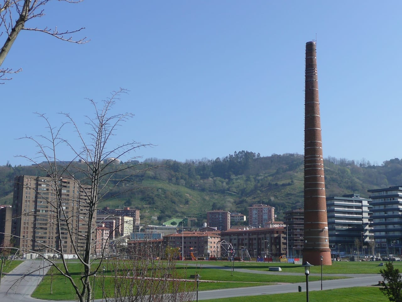 View of Parque de Etxebarria in Bilbao, featuring the iconic red brick smokestack, city buildings, green lawns, and distant hills under clear blue sky.
