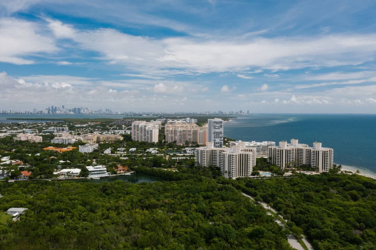 Aerial view of Miami with downtown skyline in the distance, coastal high-rises, and green space, suggesting travel west from the city.