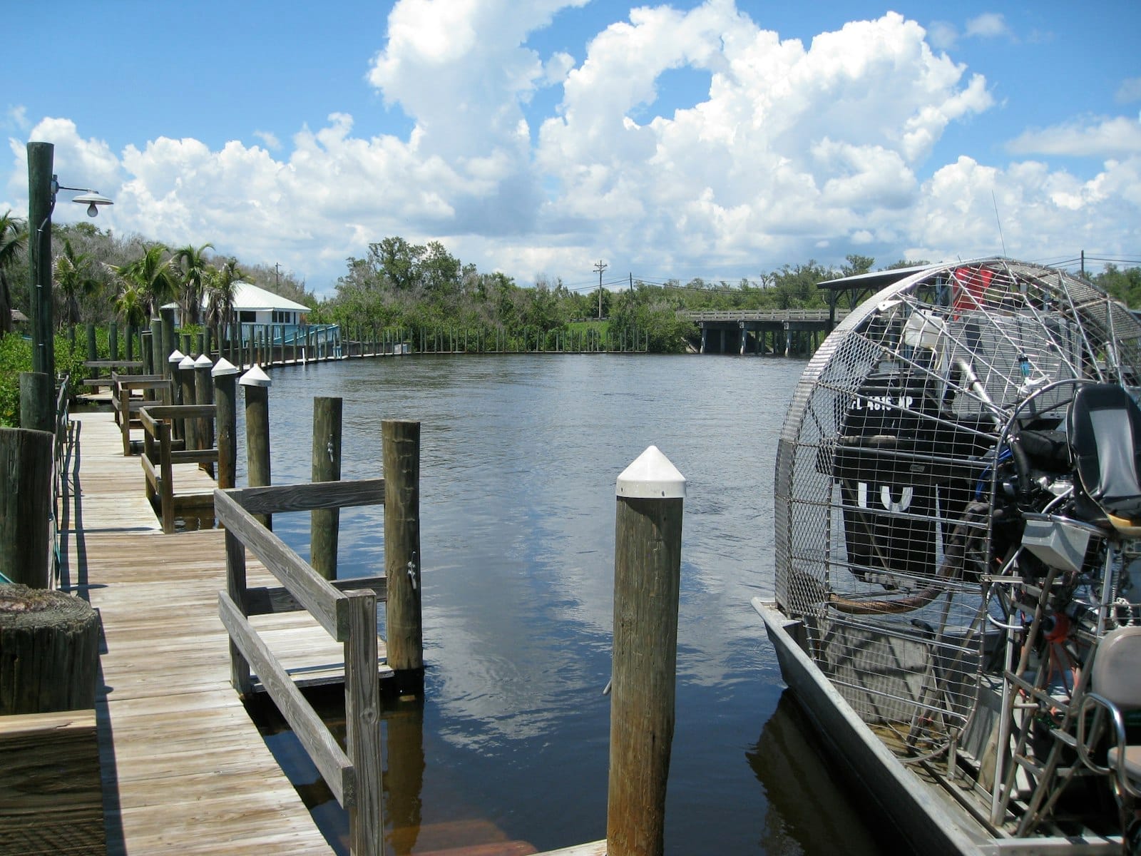 Um airboat clássico dos Everglades atracado ao lado de um píer de madeira sob céu azul, com vegetação exuberante e água calma refletindo as nuvens.