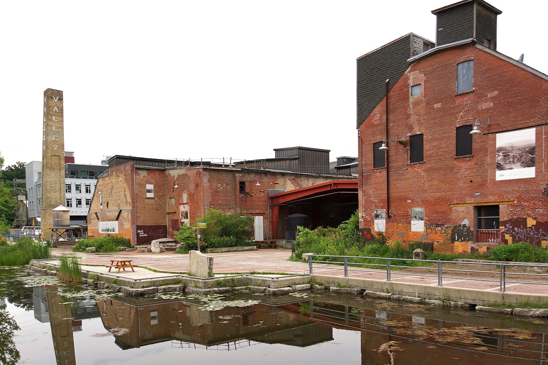 อาคารอุตสาหกรรมอิฐแดงและปล่องควันสูงสะท้อนอยู่บนผิวบึงอันสงบที่ Evergreen Brick Works ในวันที่ท้องฟ้าครึ้ม
