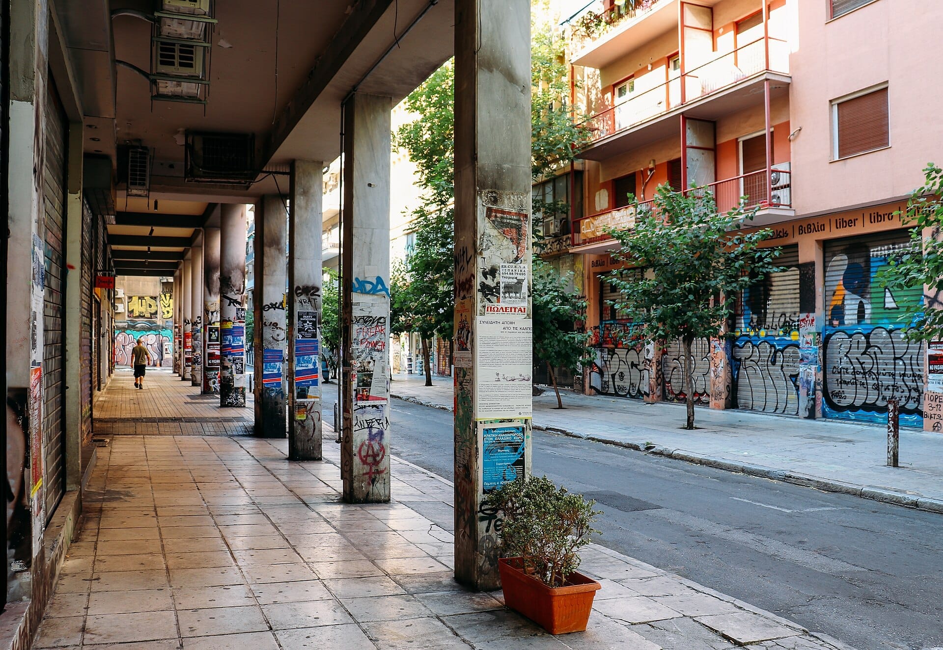ภาพถนนในย่าน Exarchia กรุง Athens มีอาคารที่เต็มไปด้วยกราฟฟิตี เสาคอนกรีตที่ติดโปสเตอร์เต็มไปหมด และคนเดินถนนคนเดียวที่กำลังเดินอยู่ใต้ซุ้มโค้ง