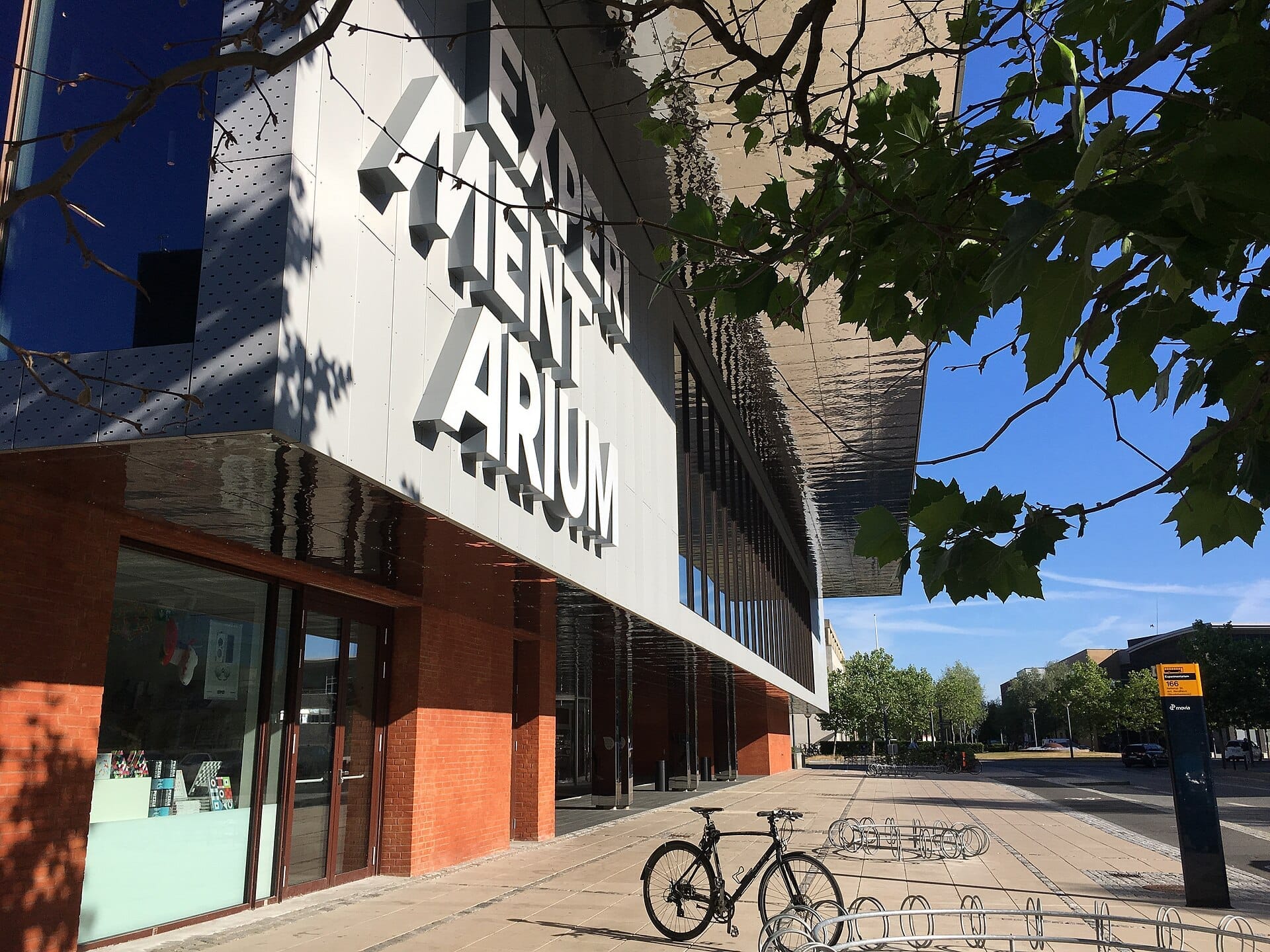 Ingresso moderno dell'Experimentarium Copenhagen con grandi insegne, facciata in mattoni rossi, finestre in vetro e una bicicletta parcheggiata fuori in una giornata di sole.