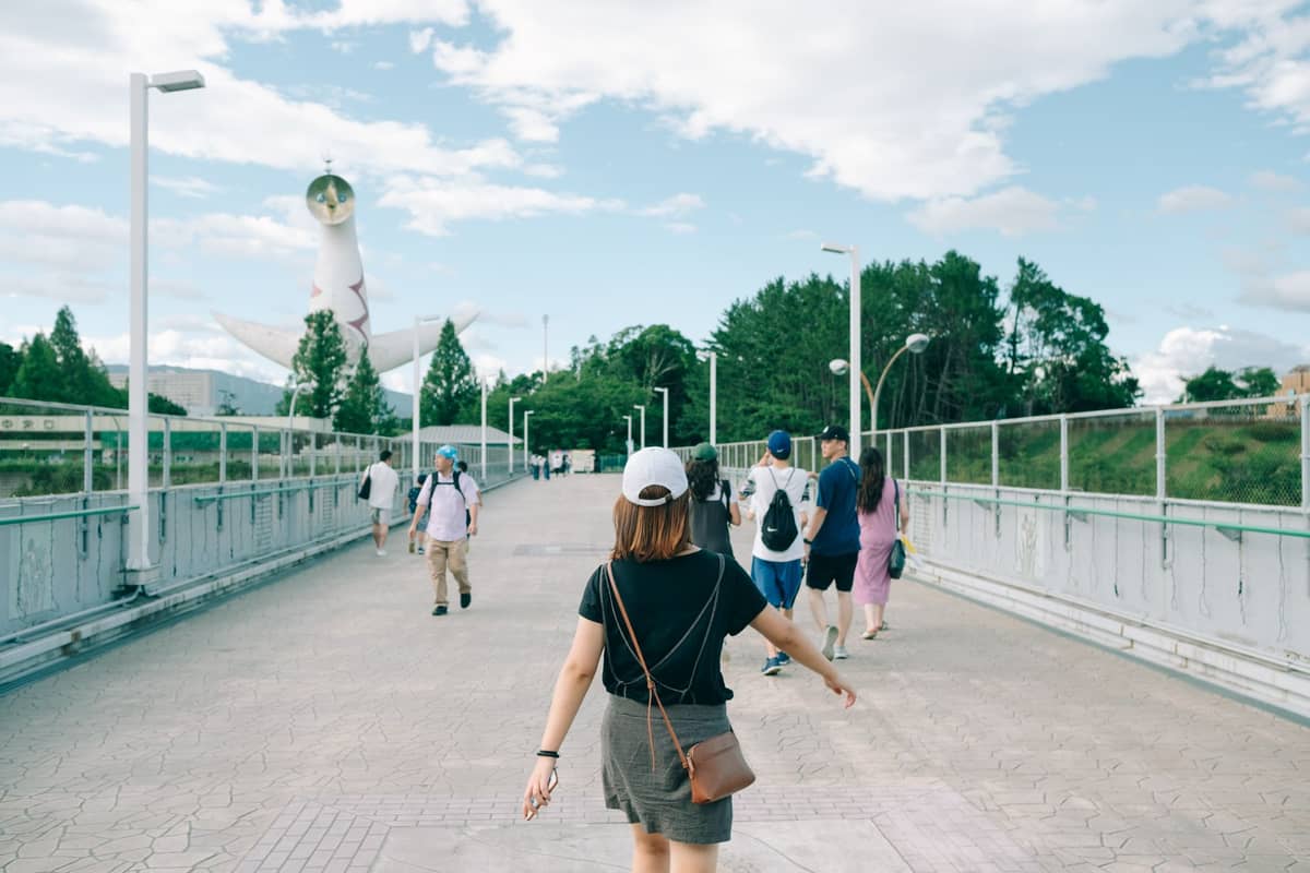 People walking toward the Tower of the Sun statue on a wide bridge at Expo '70 Commemorative Park in Osaka, with lush greenery and blue sky.