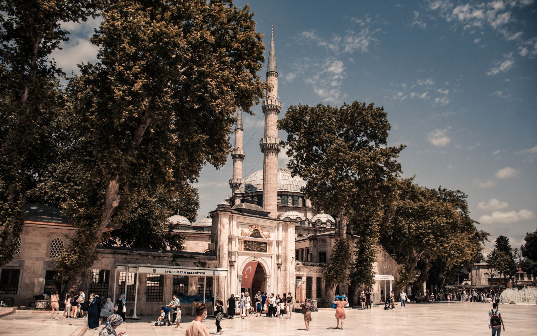 La Moschea di Eyüp Sultan con il suo imponente ingresso e i minareti, circondata da alberi alti e una piazza animata piena di persone sotto un cielo luminoso.