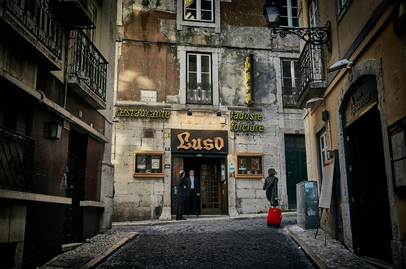 Vista da rua da entrada do Luso, um restaurante tradicional de fado em Lisboa com duas pessoas de pé do lado de fora sob luzes de néon amarelas.