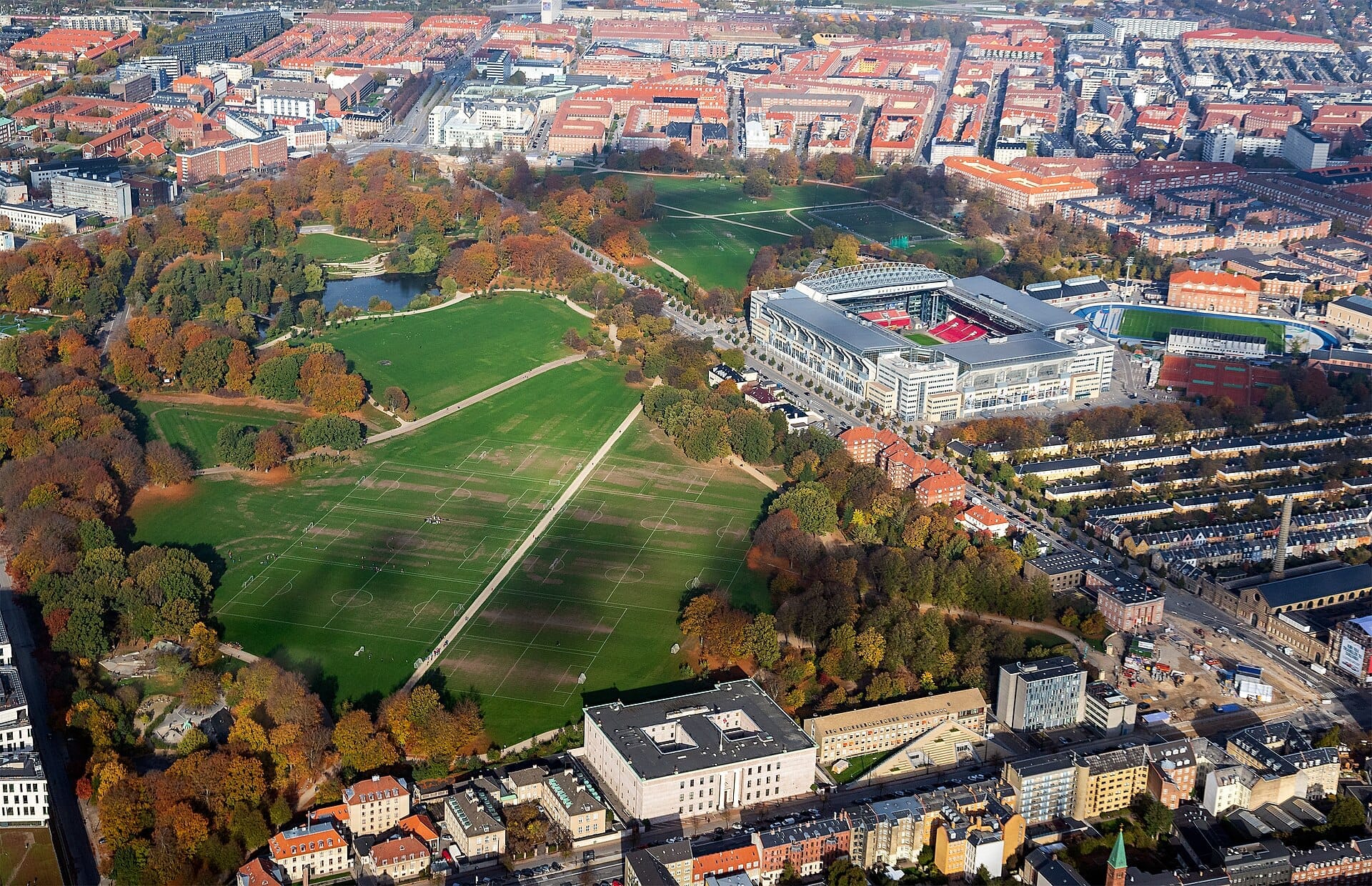 Vista aerea di Fælledparken a Copenaghen con prati verdi, vialetti alberati, campi sportivi, uno stagno e un grande stadio moderno nelle vicinanze.
