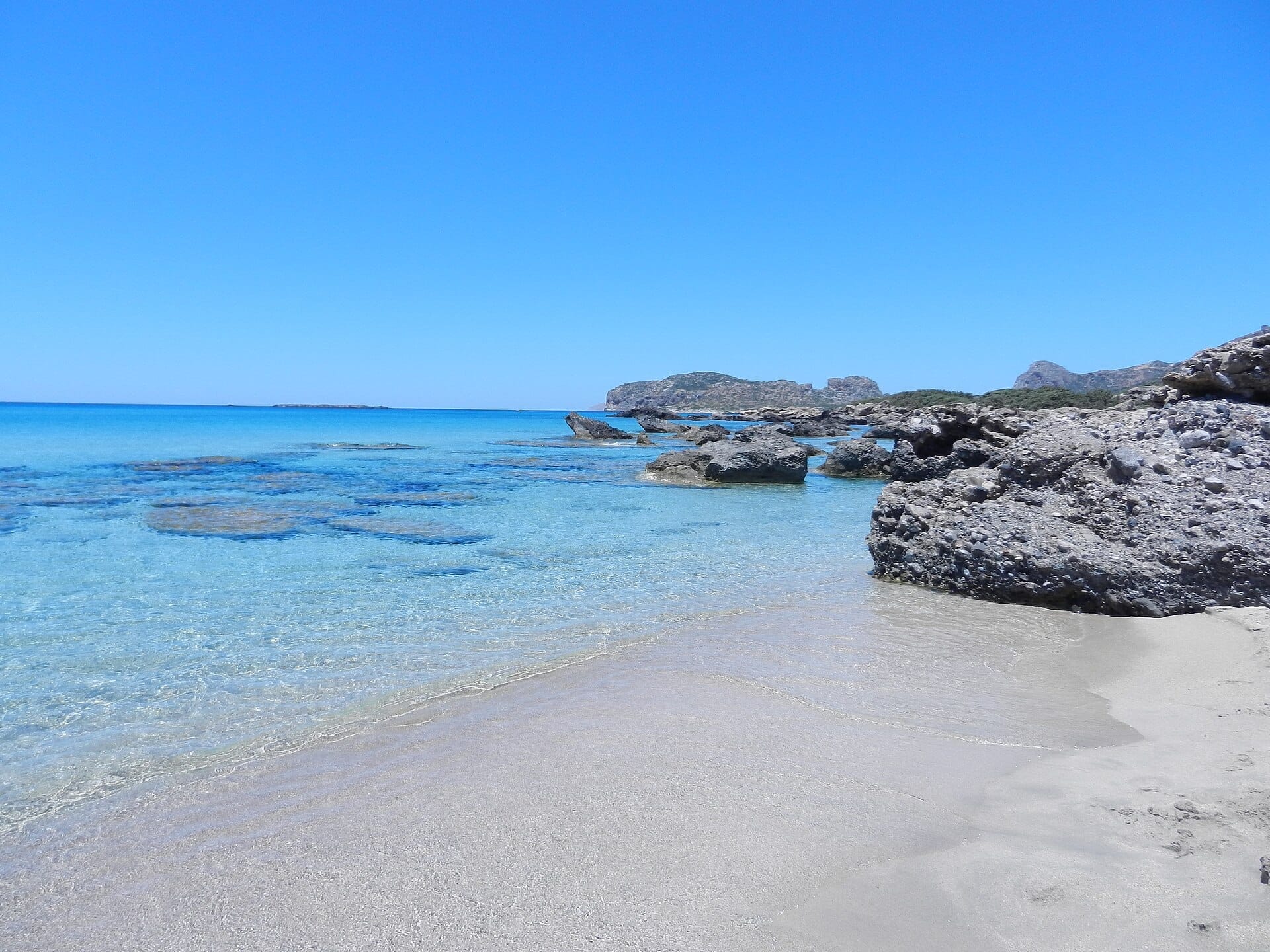 Acque turchesi cristalline e sabbia chiara a Falassarna Beach, Creta, con affioramenti rocciosi sparsi e colline lontane sotto un cielo azzurro luminoso.