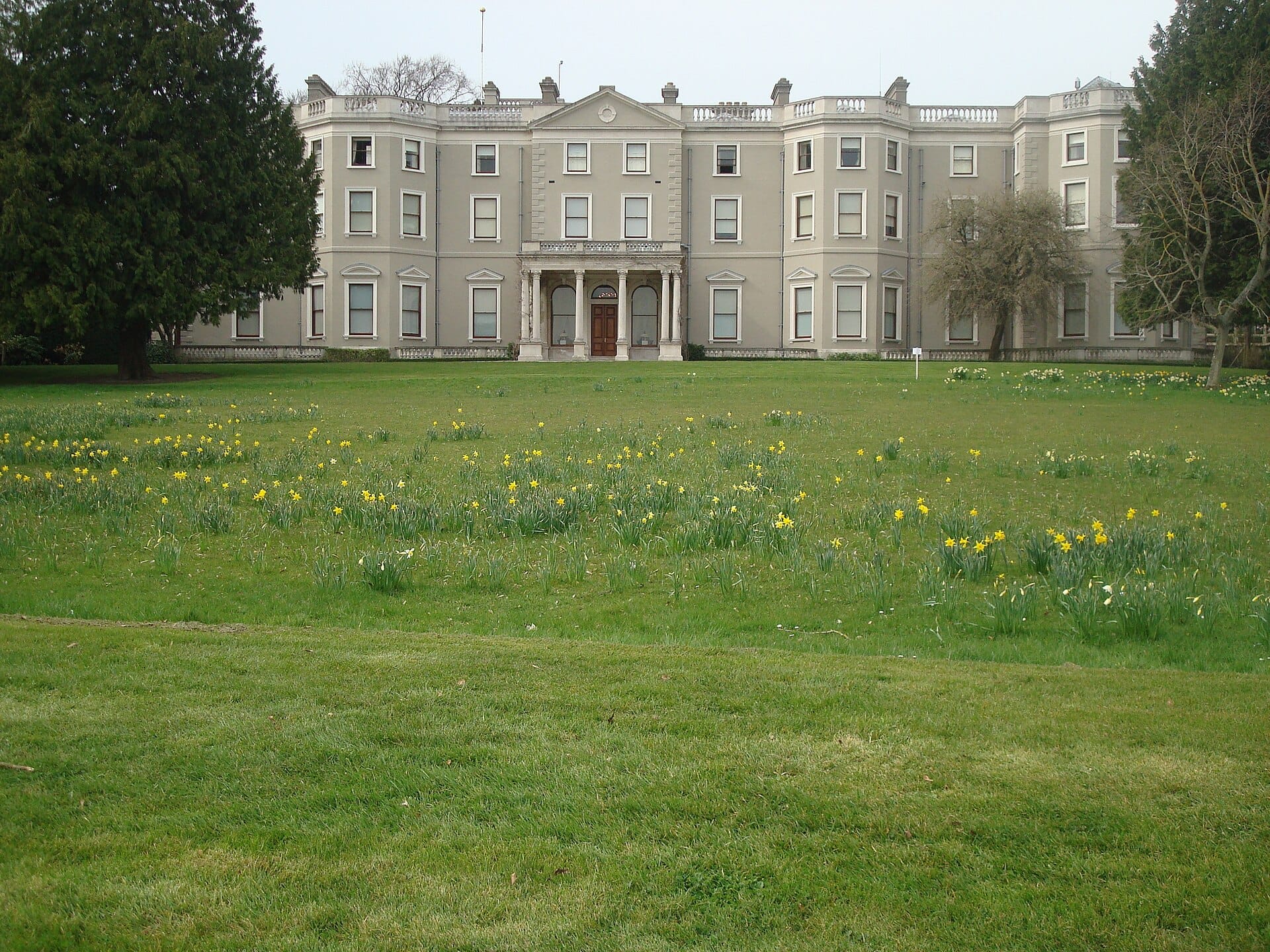 Farmleigh House situata dietro un ampio prato verde costellato di narcisi gialli, incorniciata da alberi maturi nel Phoenix Park, Dublino.