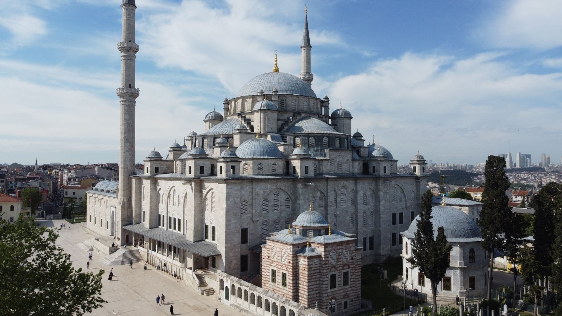 Vista esterna grandangolare della Moschea di Fatih a Istanbul sotto un cielo blu con qualche nuvola, con le cupole, i minareti e il cortile circostante dove camminano alcune persone.